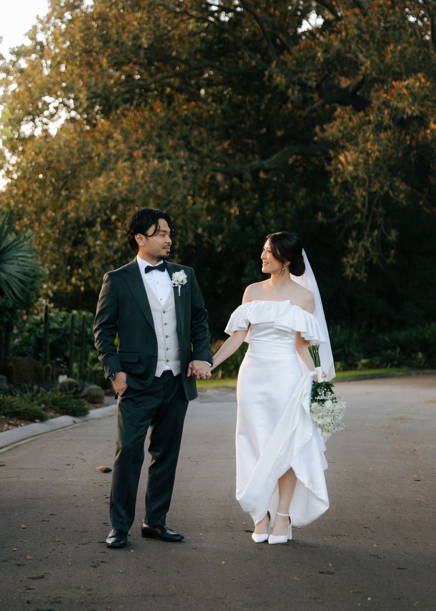 Bride and groom holding hands and smiling at each other while strolling along a tree-lined path at golden hour.