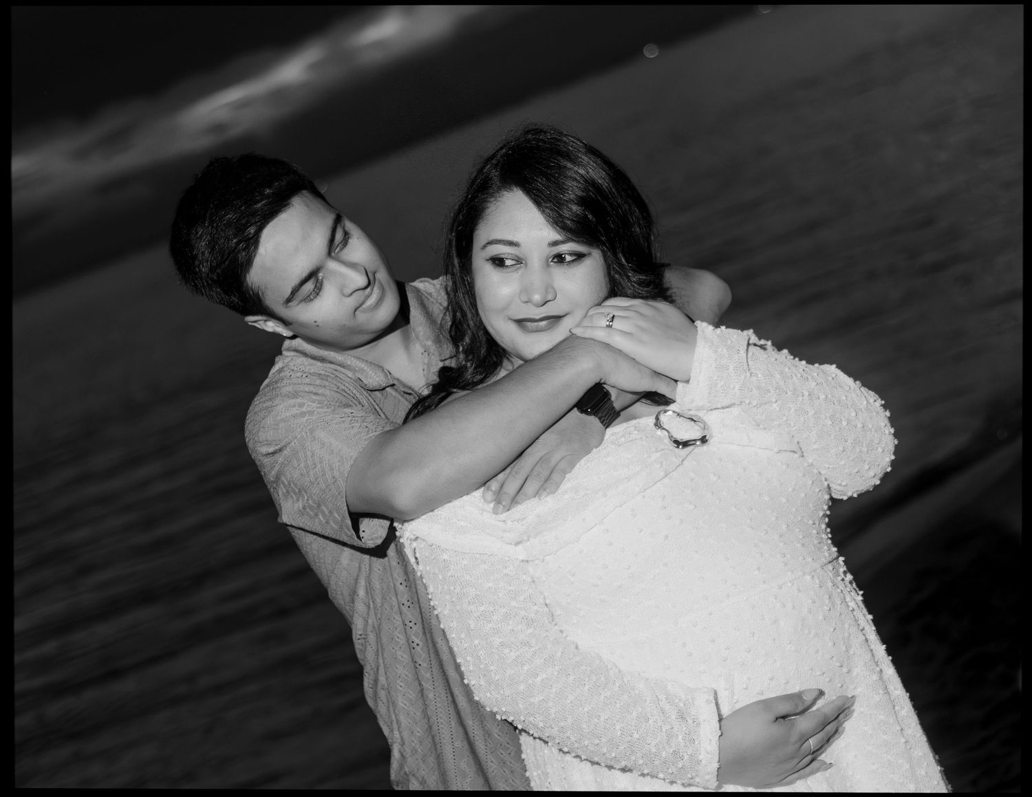 Black and white photo of couple embracing, man hugging pregnant woman from behind near water.
