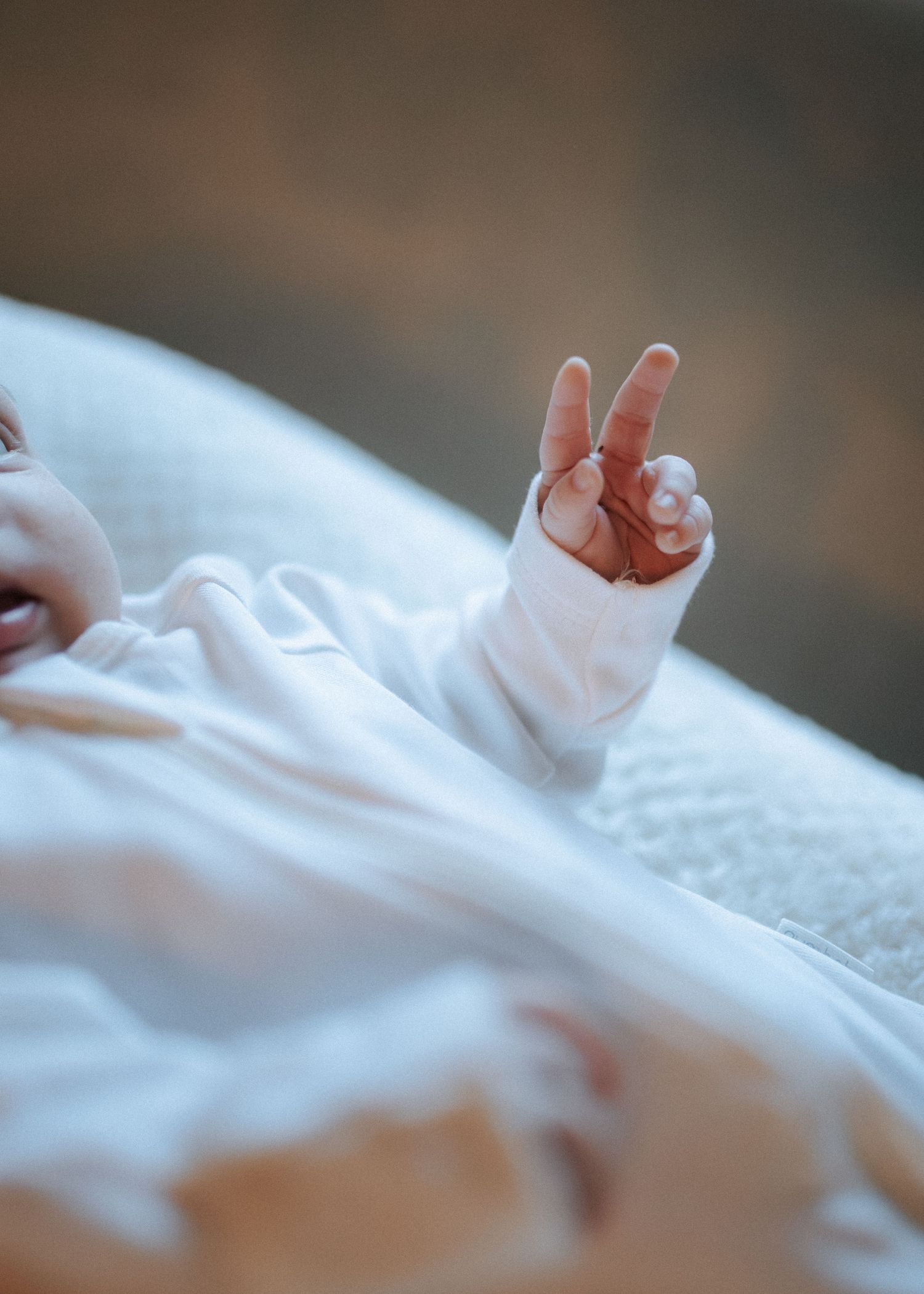Newborn baby making a peace sign with tiny fingers in white onesie.