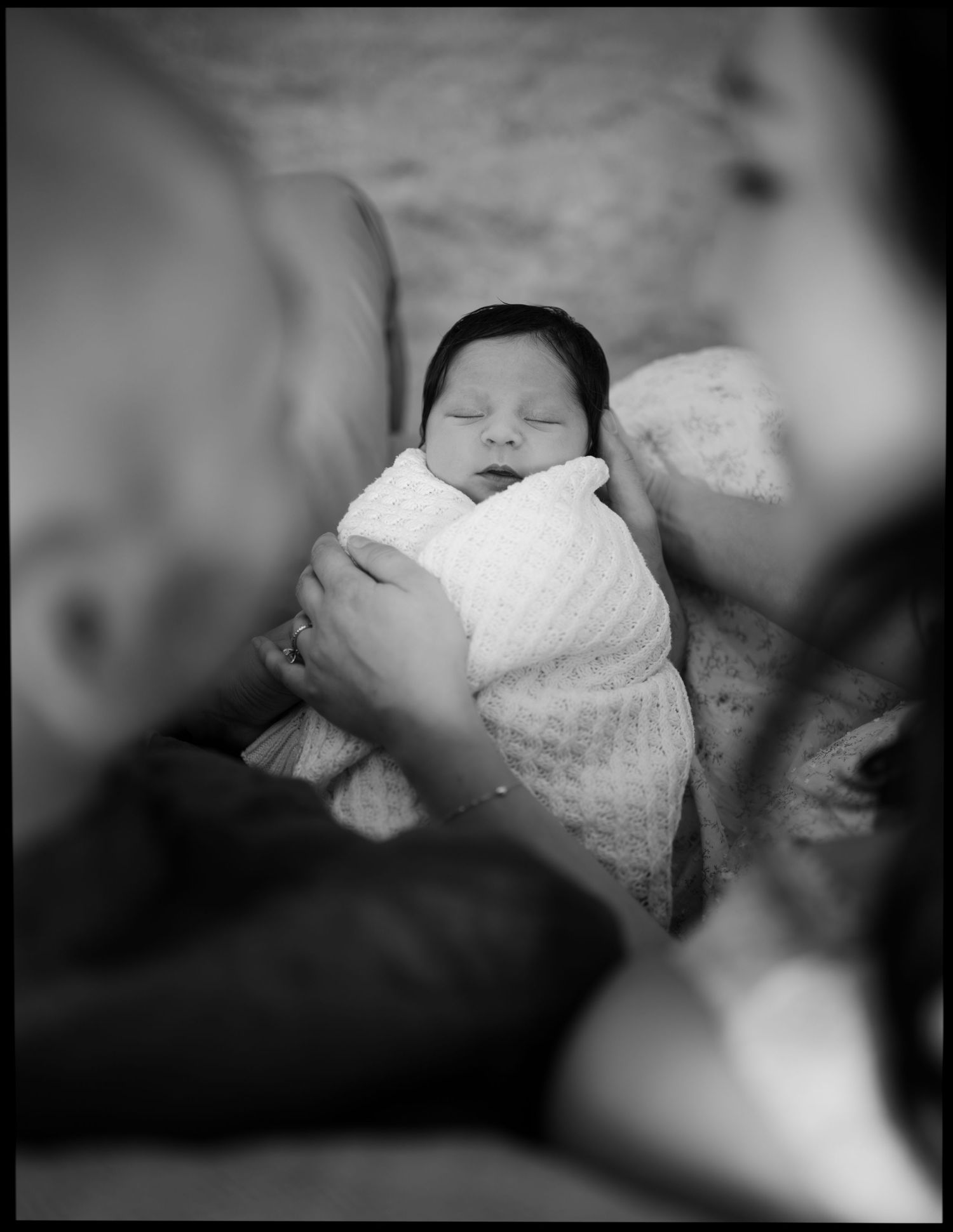 Black and white photo of sleeping newborn wrapped in white swaddle held by parents.