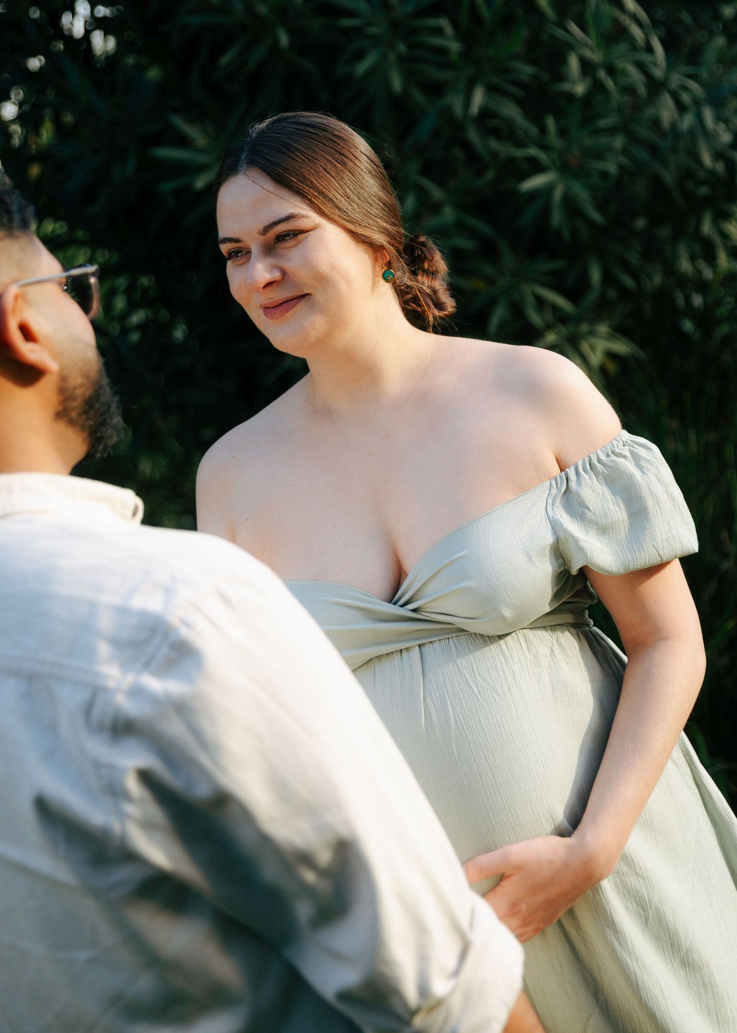 Pregnant woman in sage off-shoulder dress smiling at partner during outdoor maternity photo session at sunset.