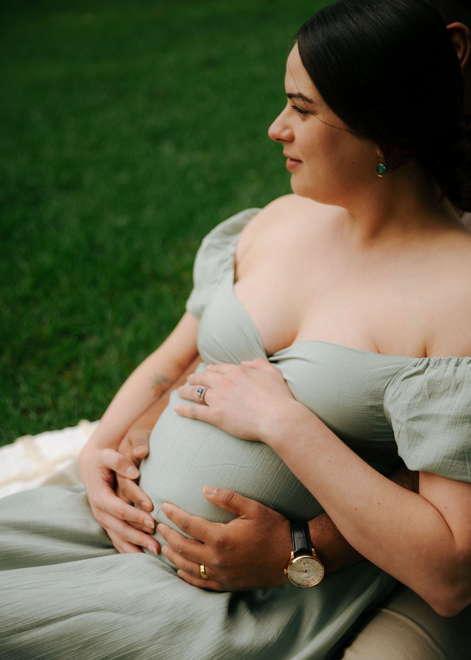 Pregnant woman in sage green off-shoulder dress cradling baby bump while sitting on grass.