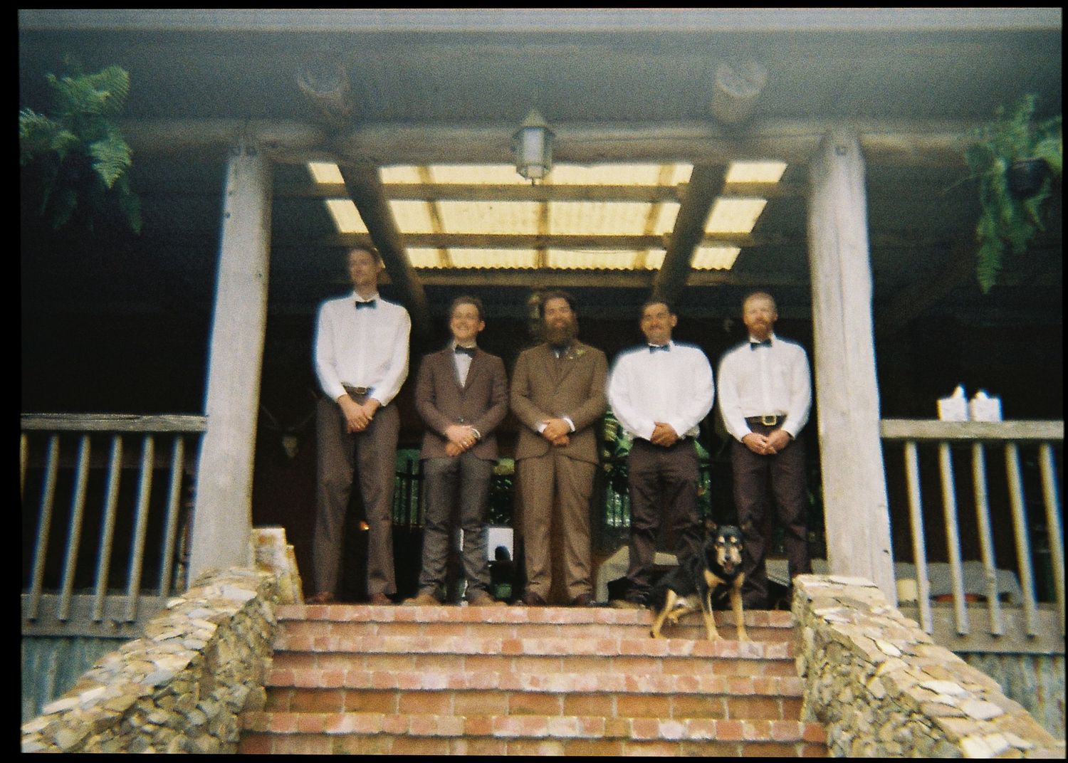 Five men in formal attire stand on stone steps of a rustic porch with wooden columns at a wedding.