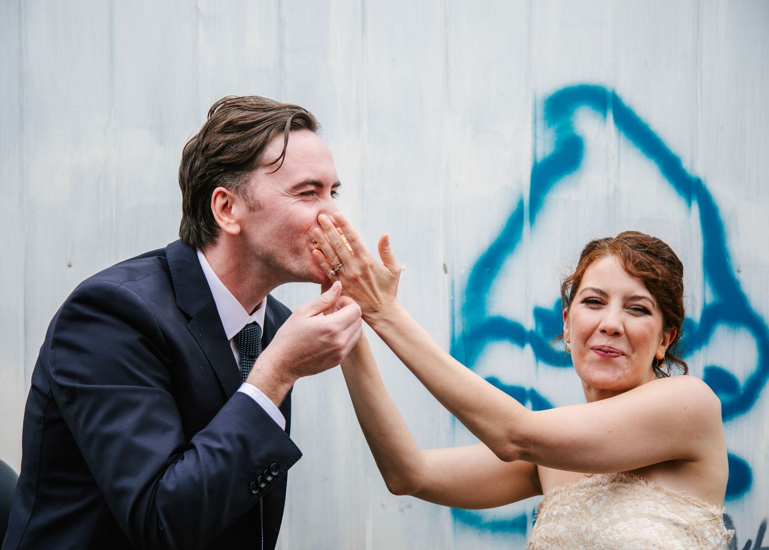 Bride and groom playfully kissing hands in front of a graffiti wall.