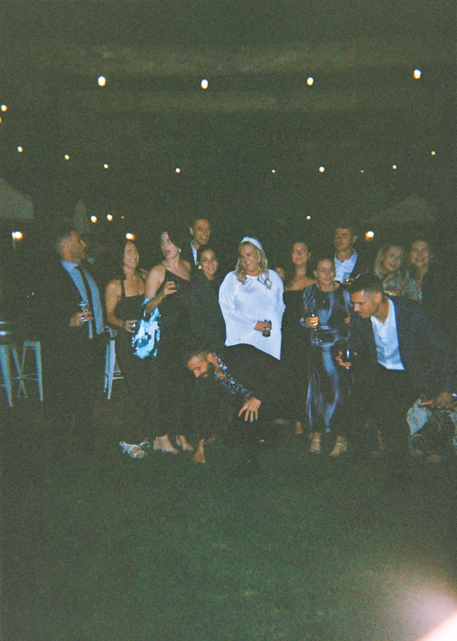 A lively group of wedding guests pose together outdoors at night under string lights during a reception celebration.