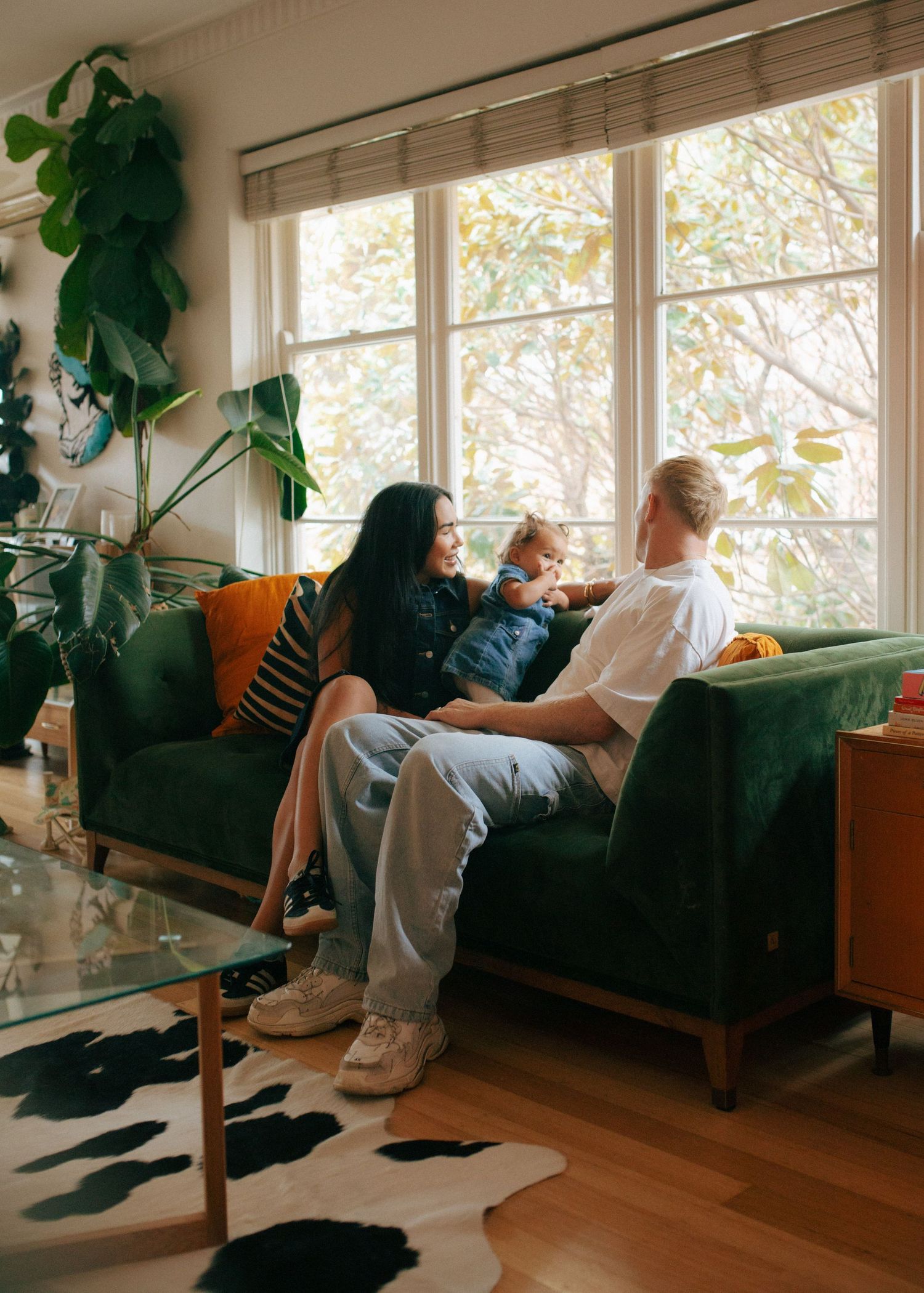 Young family relaxing on green velvet sofa in bright living room with plants and large windows.
