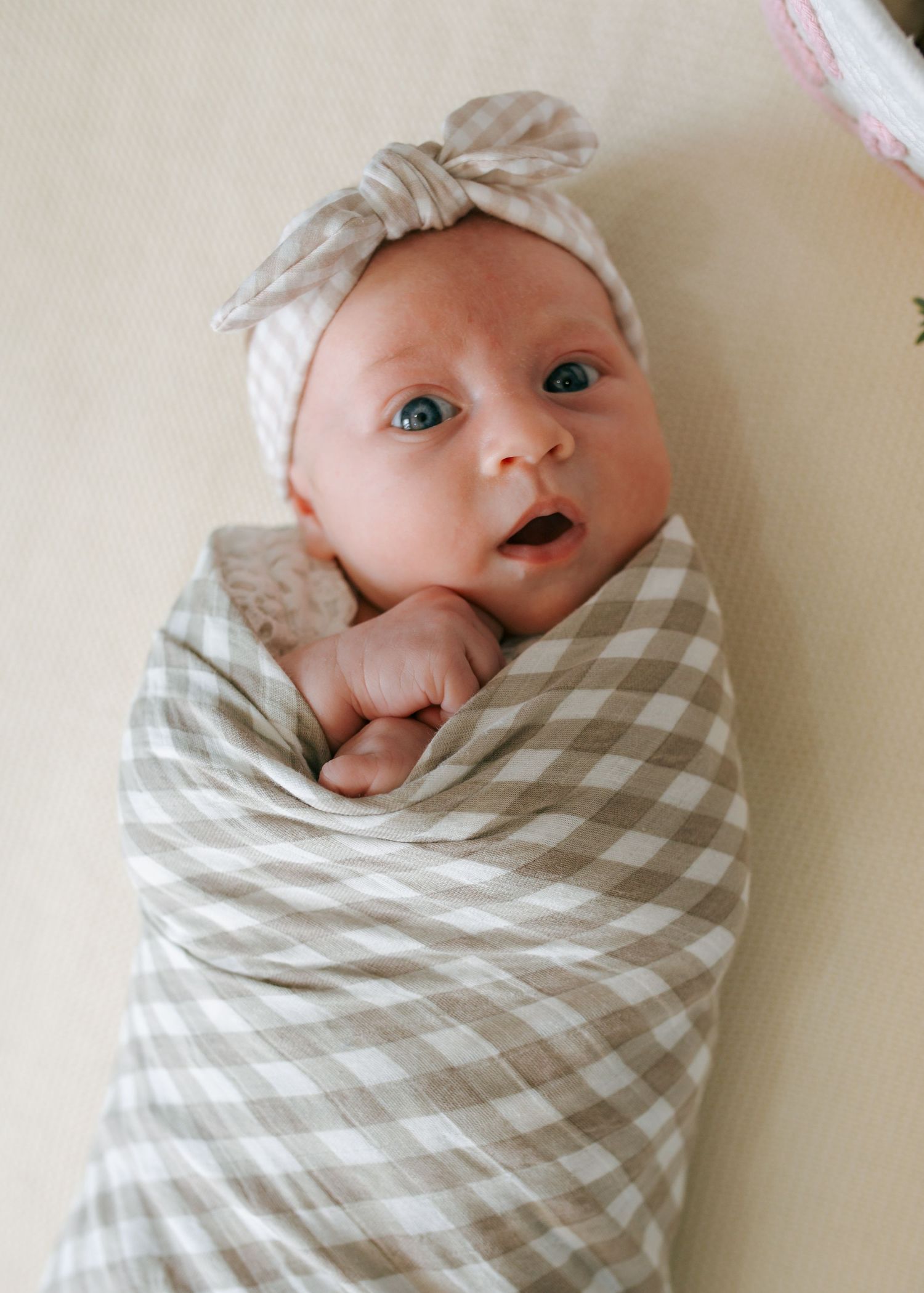 Newborn baby swaddled in tan gingham blanket with matching bow headband looking up with wide eyes.