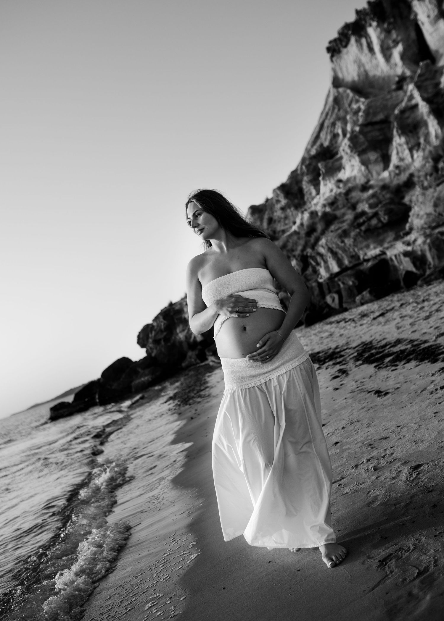 Black and white maternity portrait of pregnant woman in flowing white dress on beach at dusk.