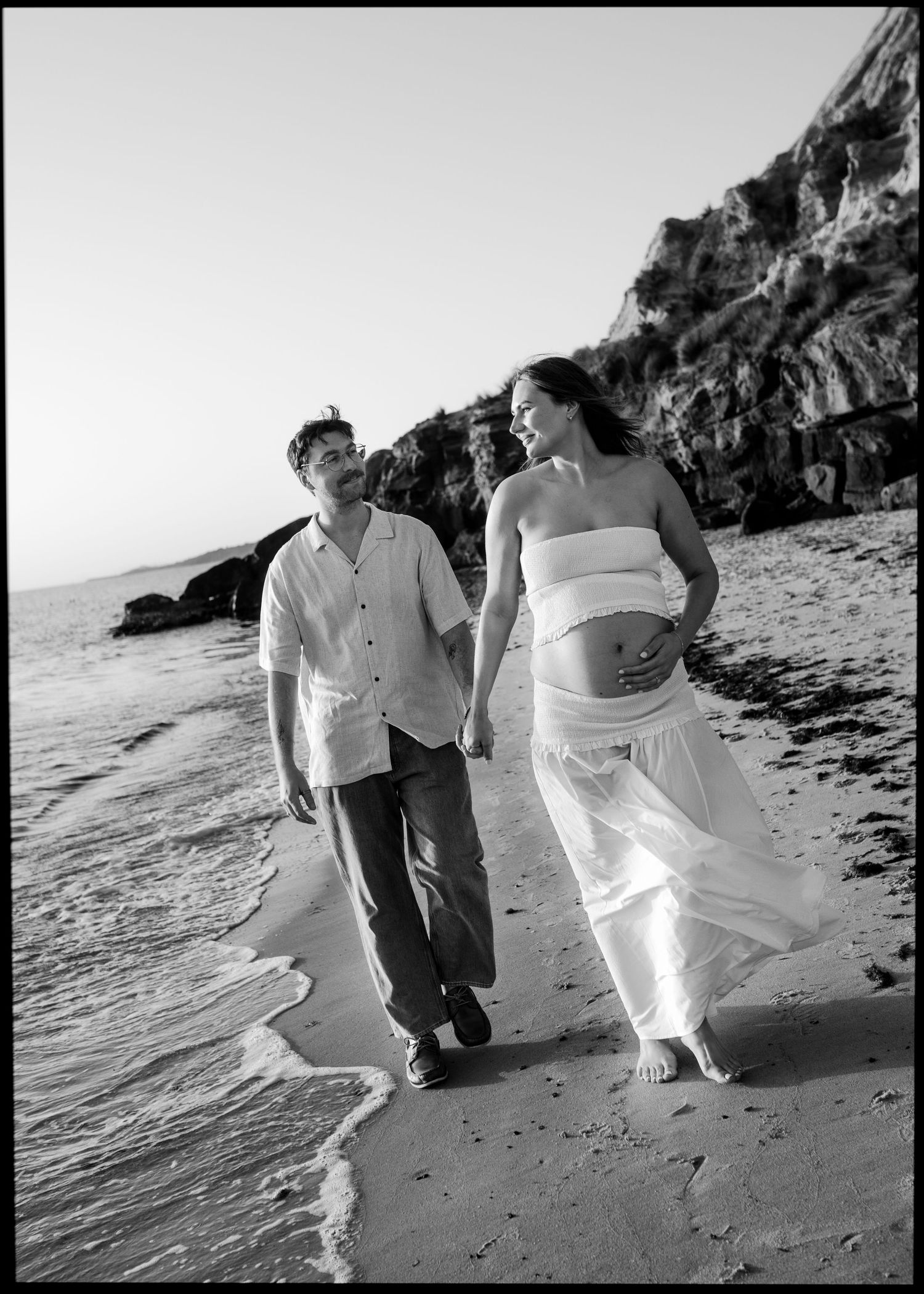 Black and white photo of pregnant couple walking hand-in-hand along beach with rocky cliffs behind.