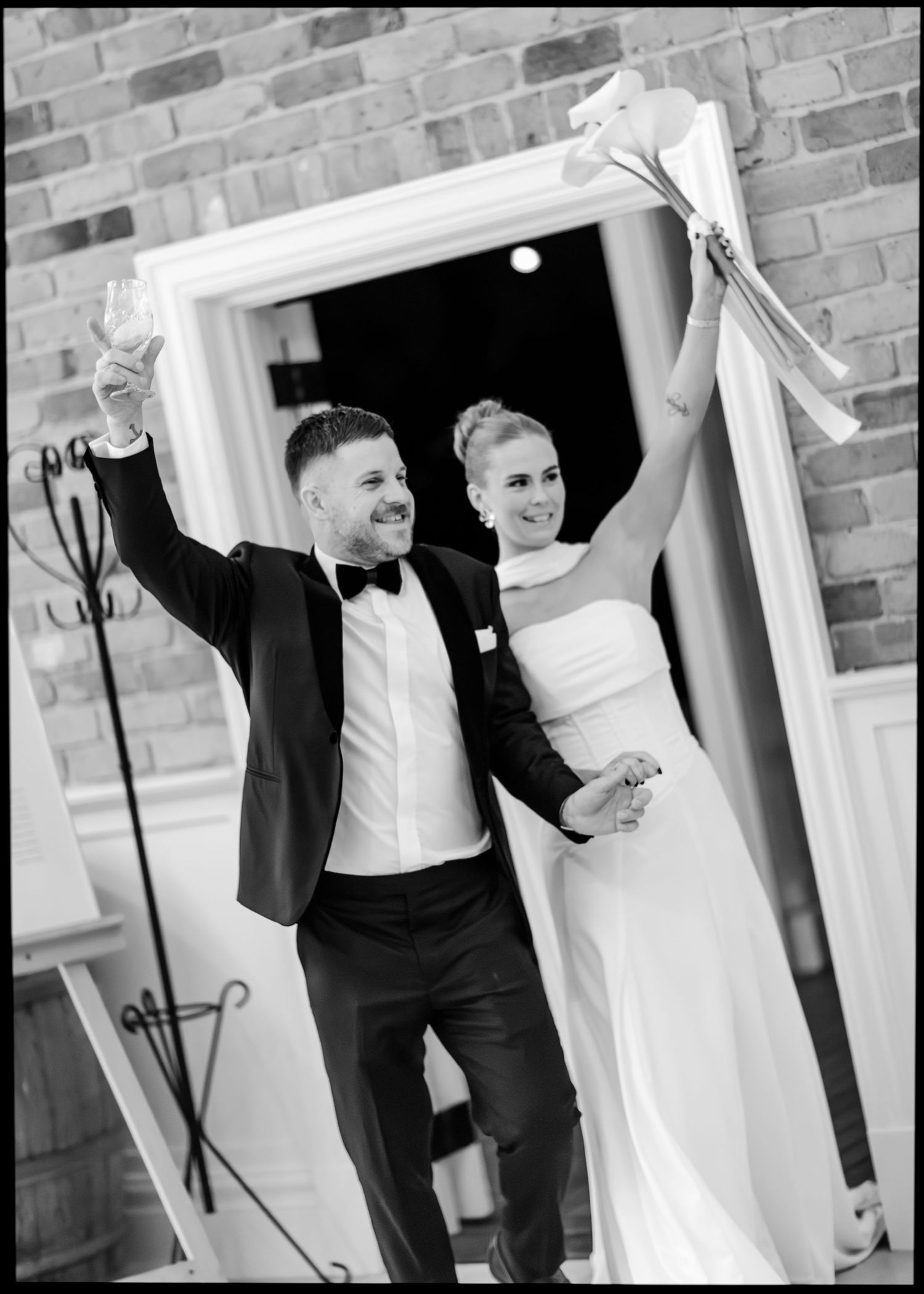 Bride and groom celebrate joyfully raising arms holding bouquet and champagne in black and white.