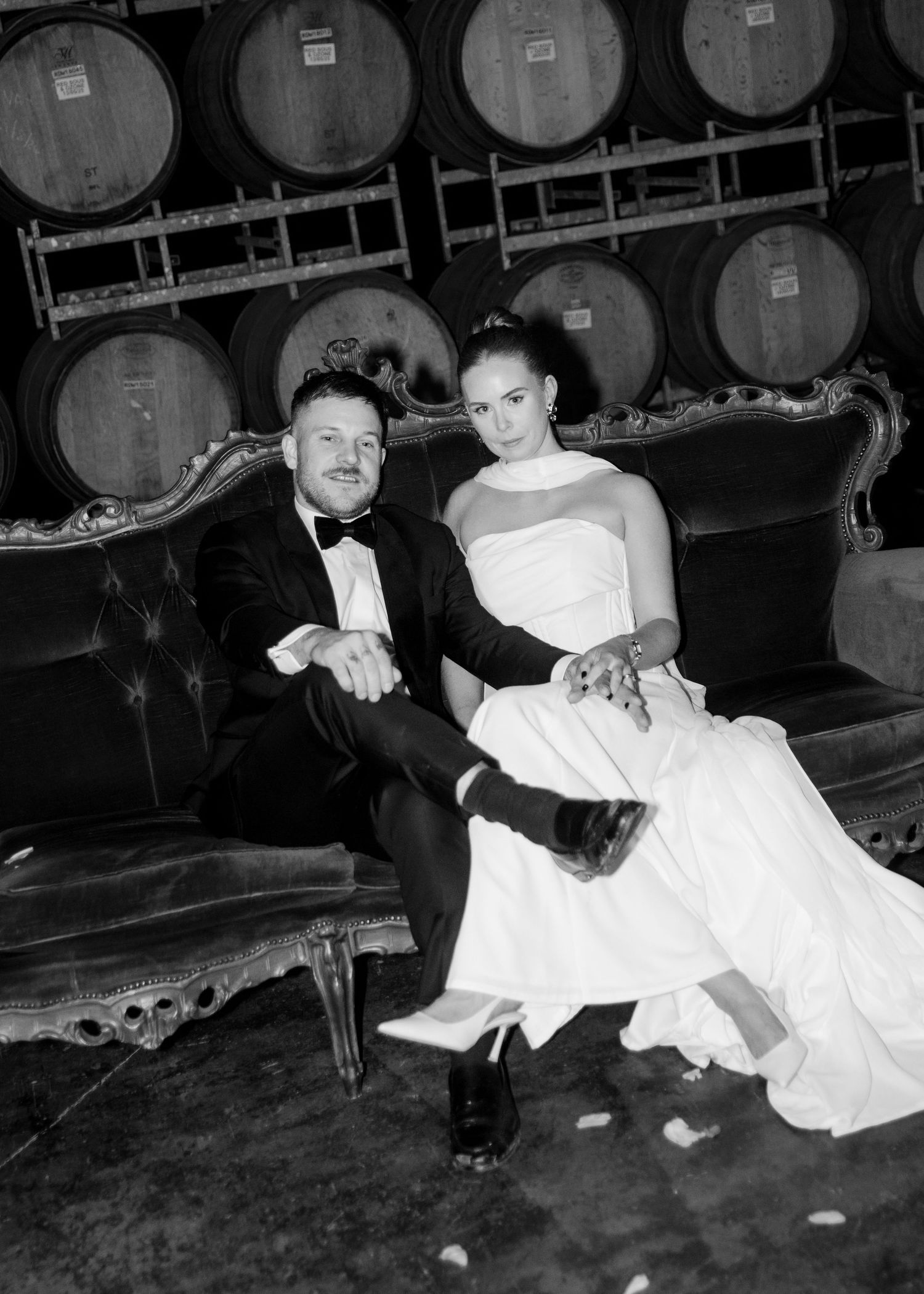 Black and white photo of a bride and groom sitting on a vintage sofa in a winery barrel room on their wedding day.