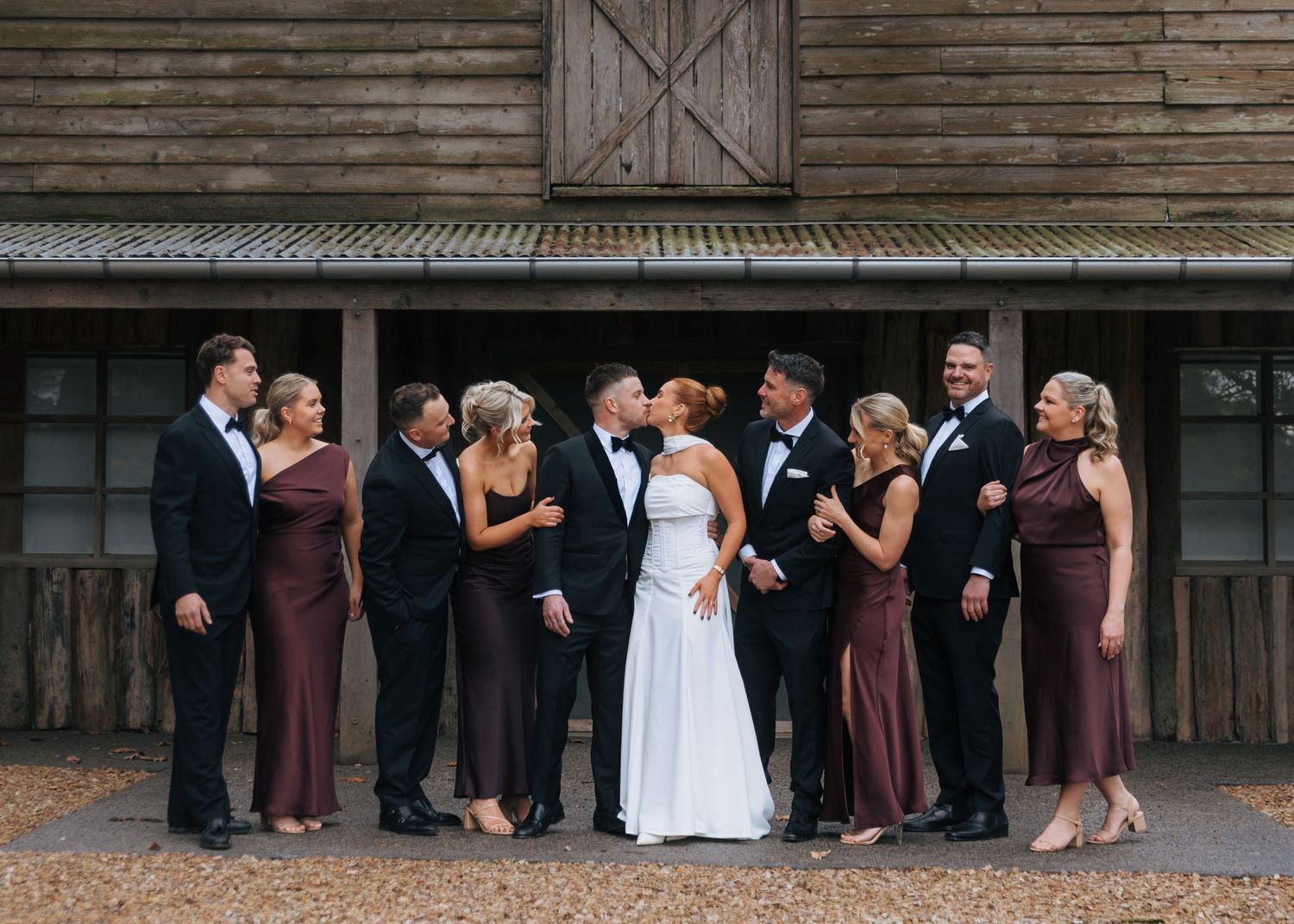 Wedding party kissing and celebrating in front of rustic barn, bridesmaids in mauve gowns and groomsmen in black tuxedos.