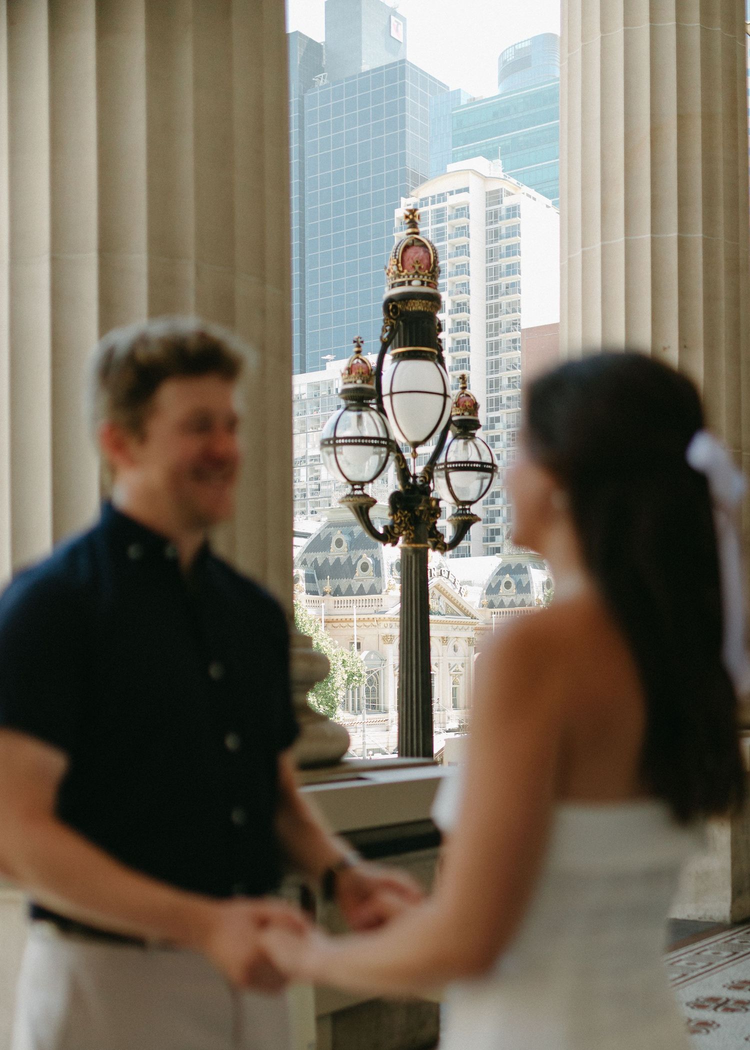 Groom smiling at bride while holding hands, with an ornate street lamp and city skyline in the background.
