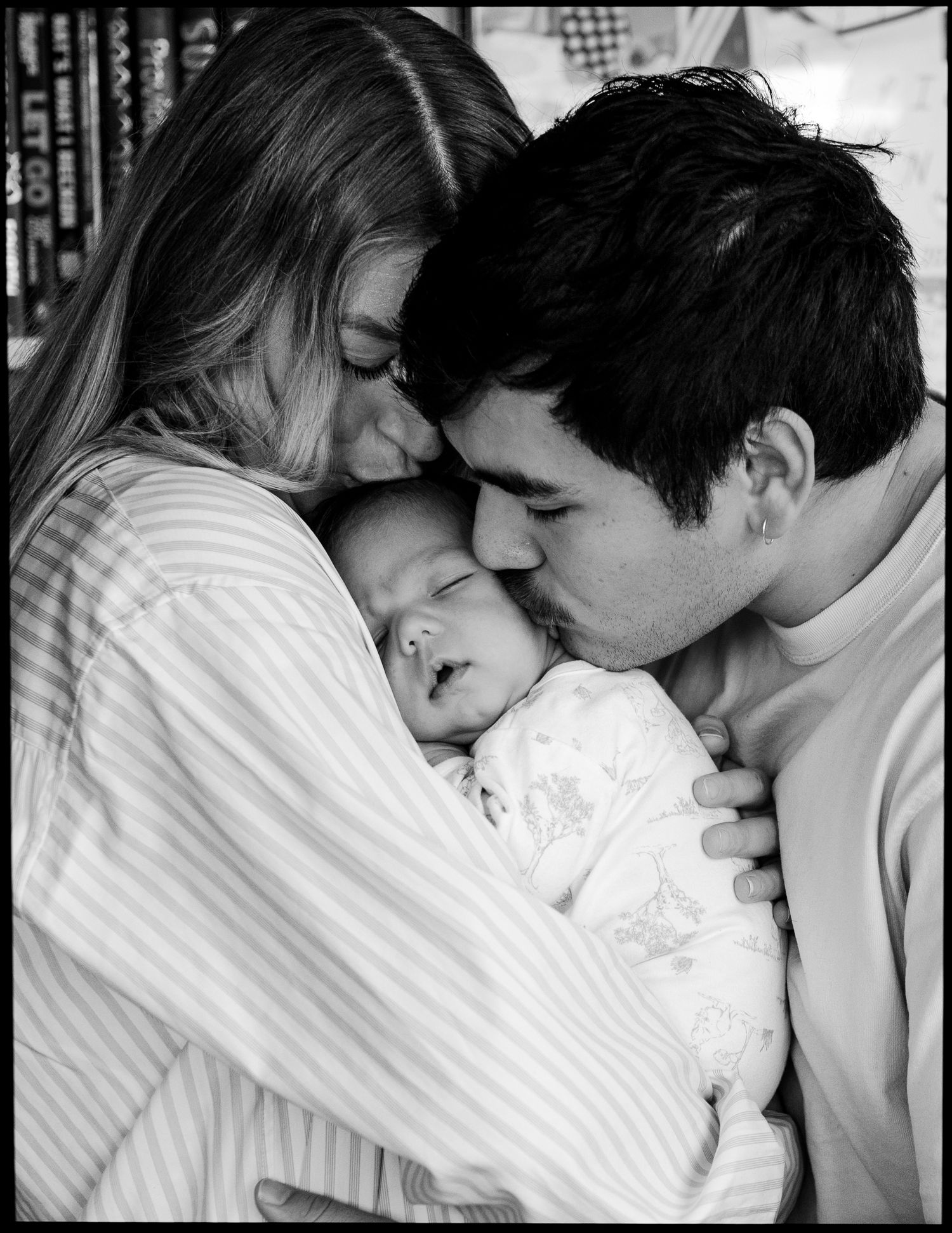 Black and white photo of two parents tenderly kissing the cheeks of their sleeping newborn baby wrapped in a soft blanket.