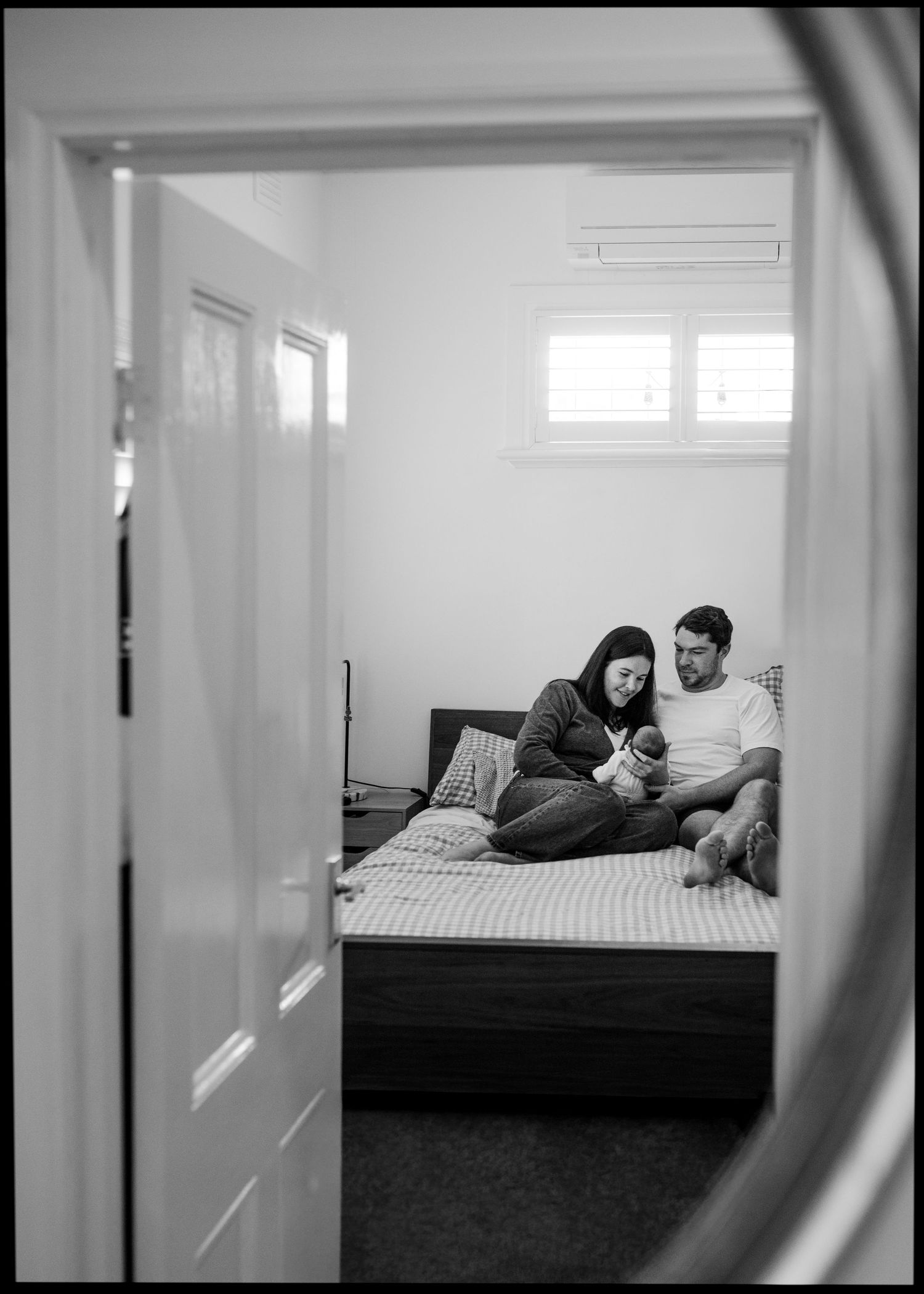 Black and white photo of new parents sitting on a bed, gently holding their newborn baby, framed through an open bedroom doorway.