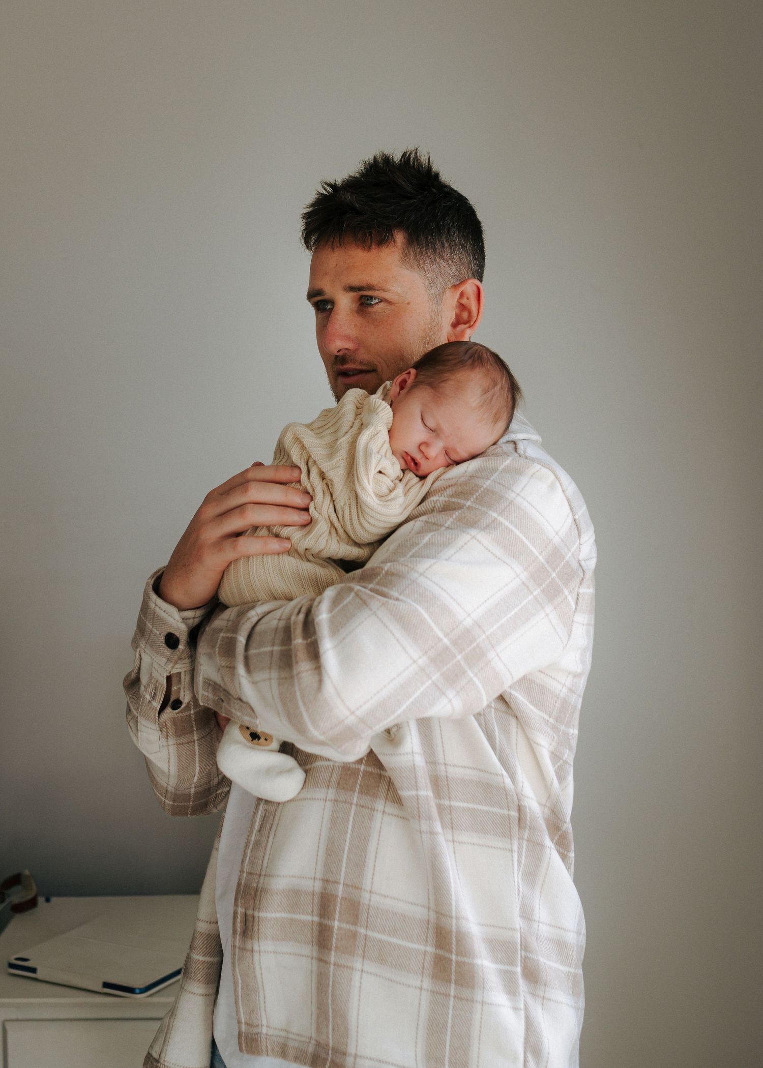 Dad in plaid shirt tenderly holding sleeping newborn baby against chest indoors.