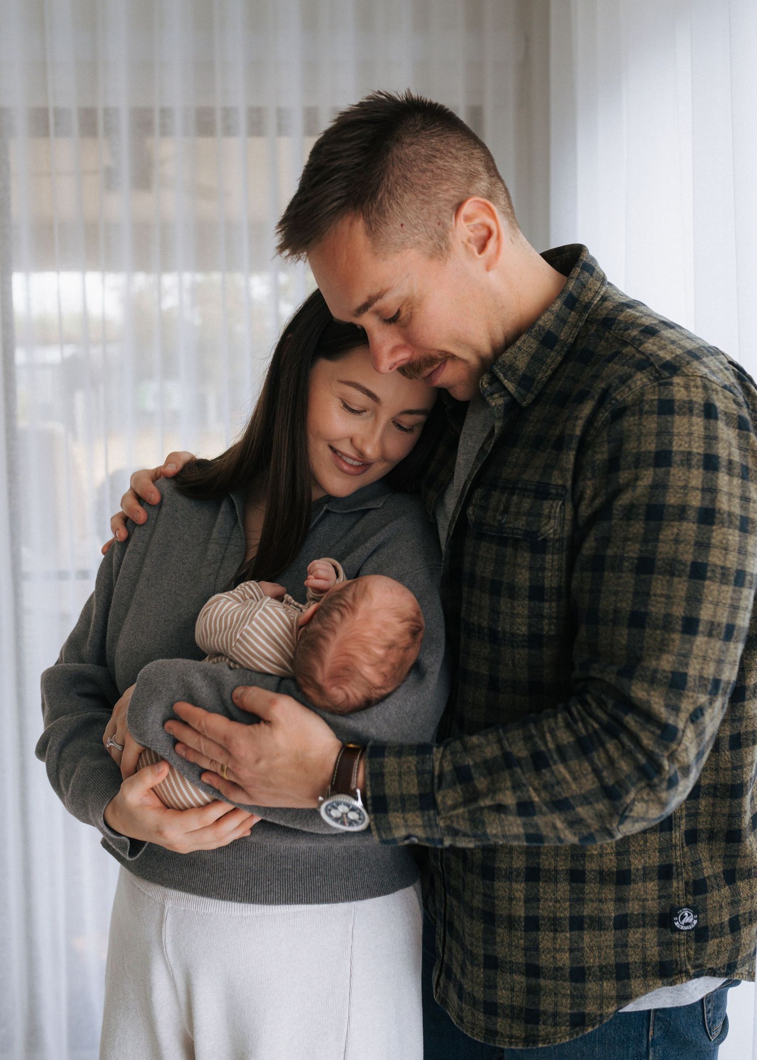 Young couple tenderly holding their newborn baby together near a bright window.