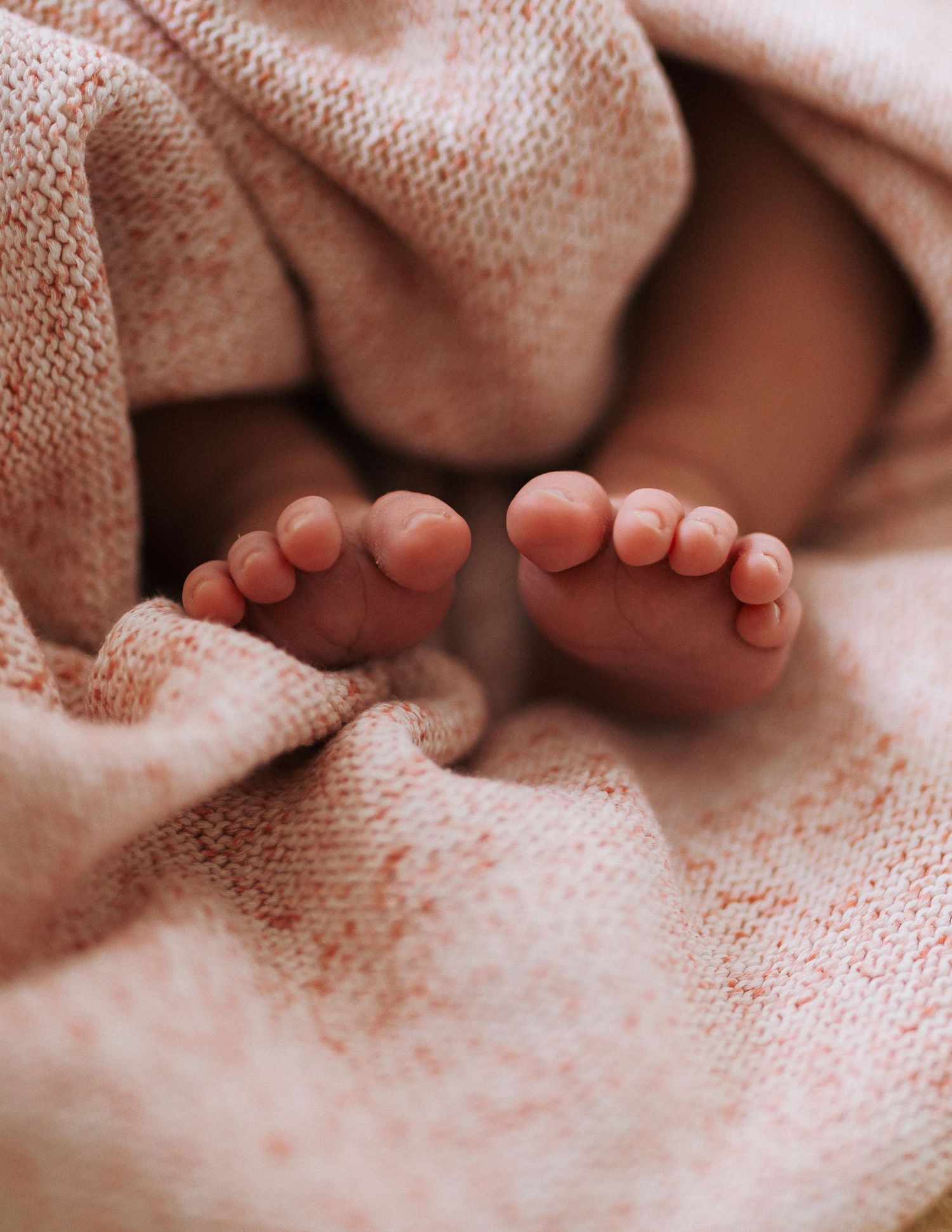 Tiny newborn baby feet peeking out from a soft, cozy peach knit blanket in warm natural light.