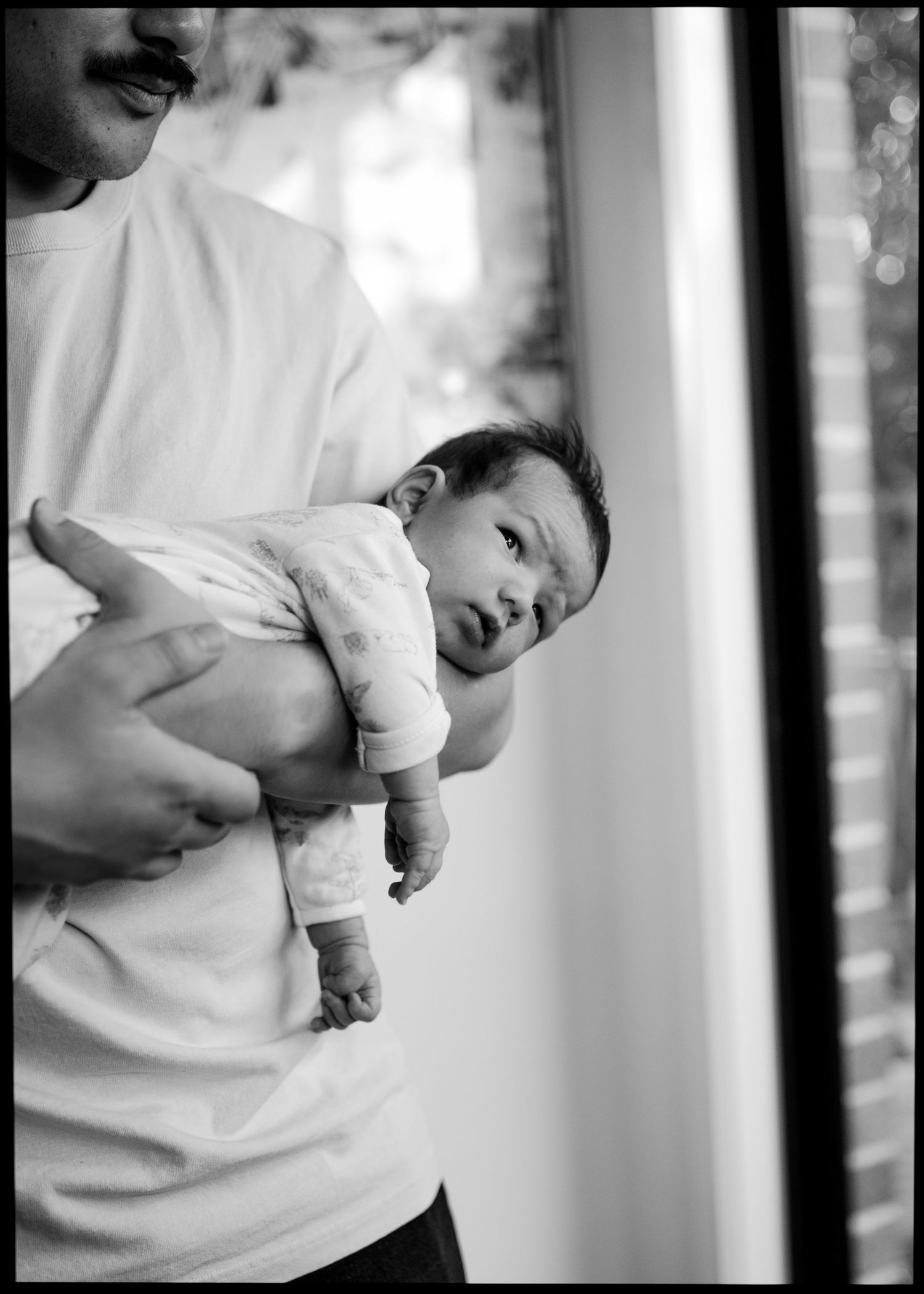 Black and white photo of a man holding a wide-eyed newborn baby against his chest while the baby gazes sideways near a window.