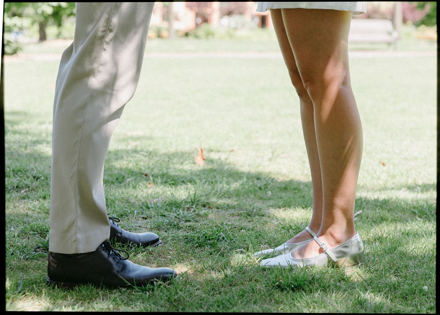 Close-up of bride and groom's shoes standing face-to-face on green grass, silver flats and black dress shoes.