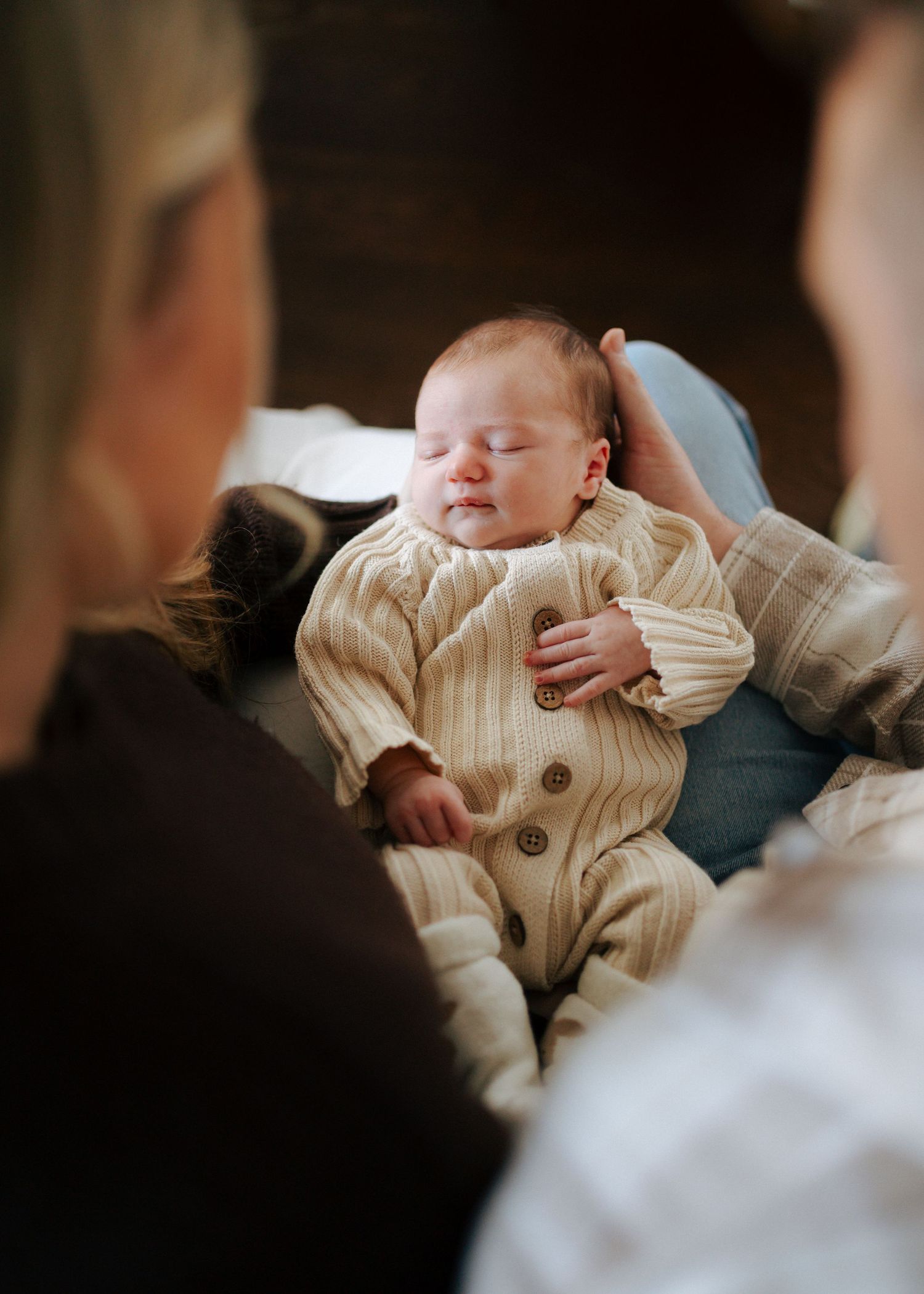 Sleeping newborn in cream ribbed onesie cradled by parents looking down lovingly.
