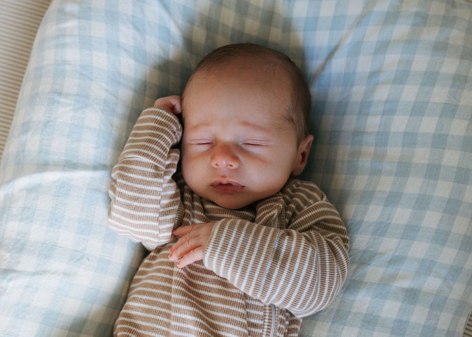 Newborn baby in tan striped pajamas resting with hand near face in blue lounger.