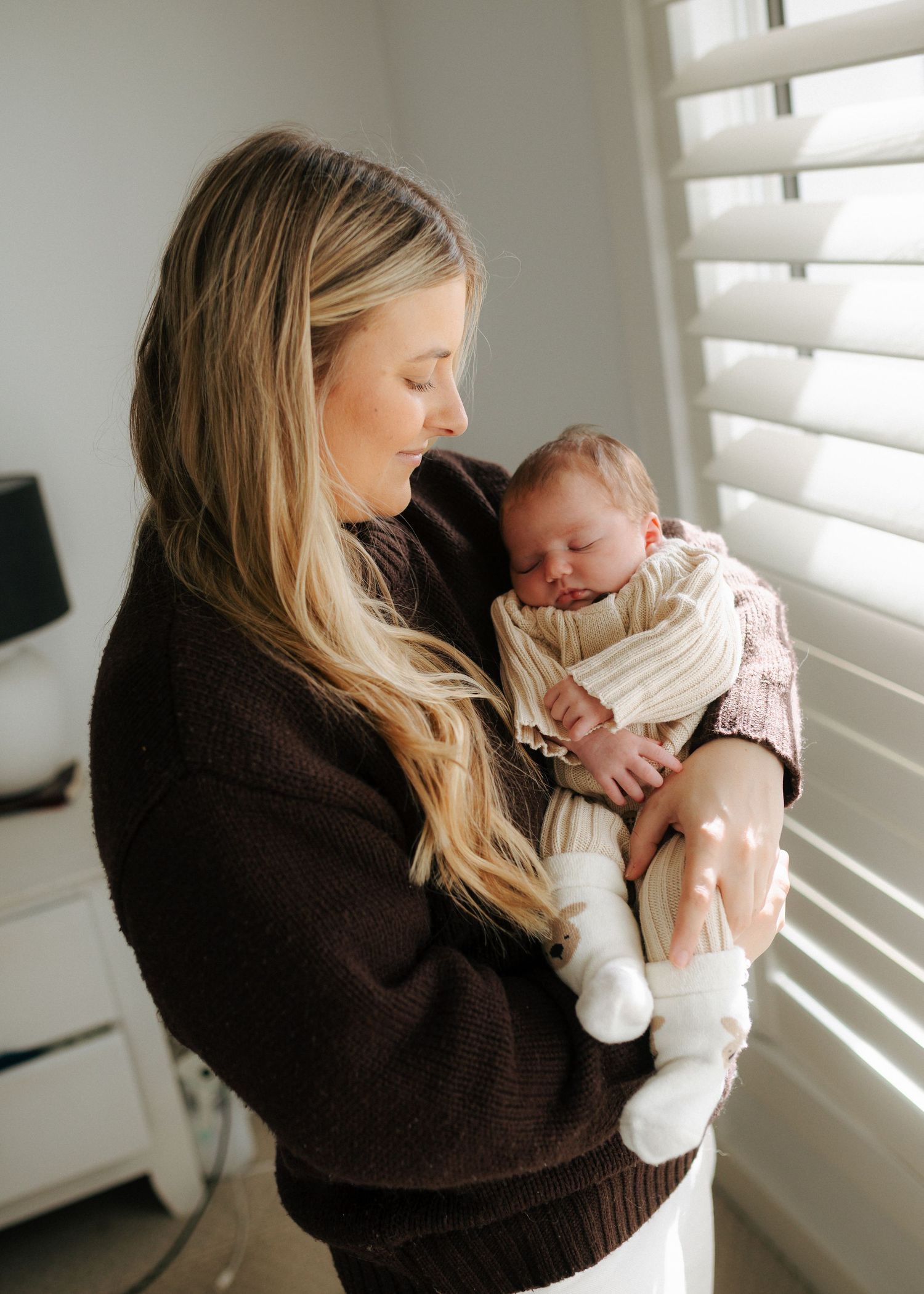 Mother in dark brown sweater cradling sleeping newborn baby near bright window with white shutters.