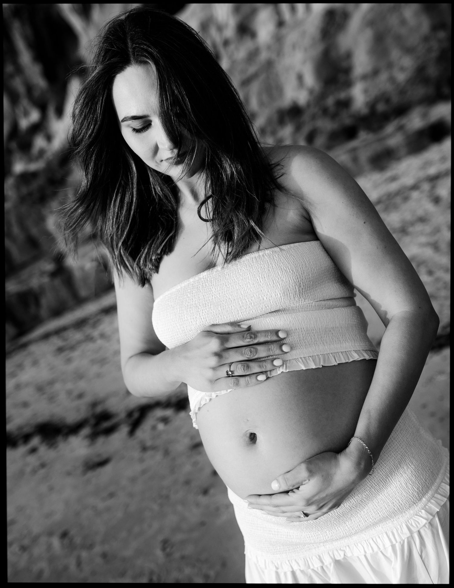 Black and white maternity photo of a pregnant woman in a white tube top cradling her baby bump outdoors.