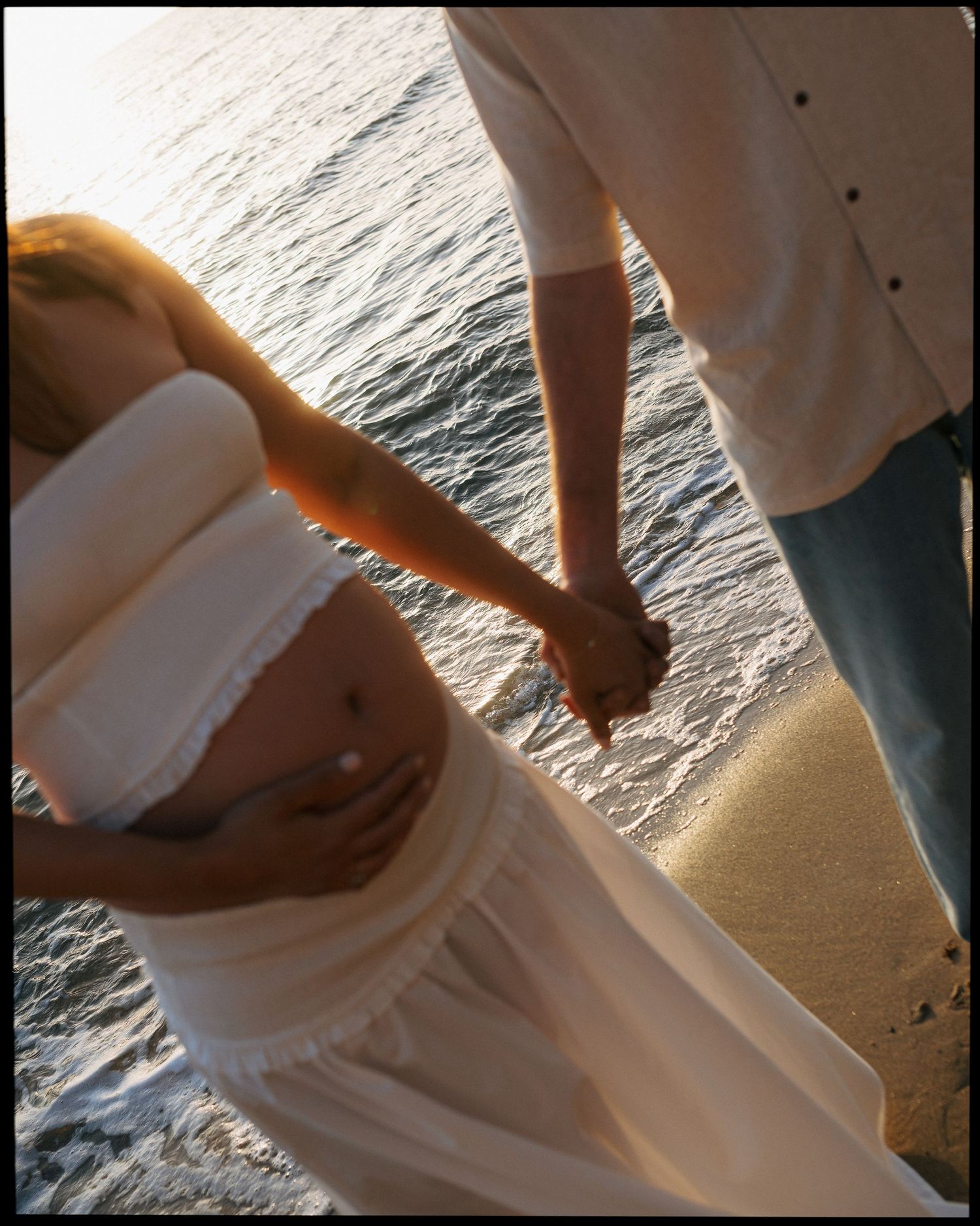 Pregnant woman in white dress holding hands with partner on sandy beach at golden sunset.