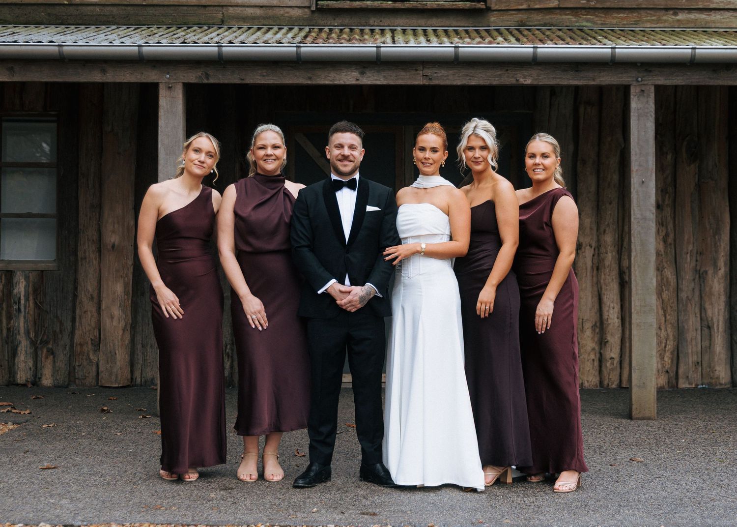 Bride in white gown and groom in tuxedo pose with four bridesmaids in burgundy dresses outside rustic venue.