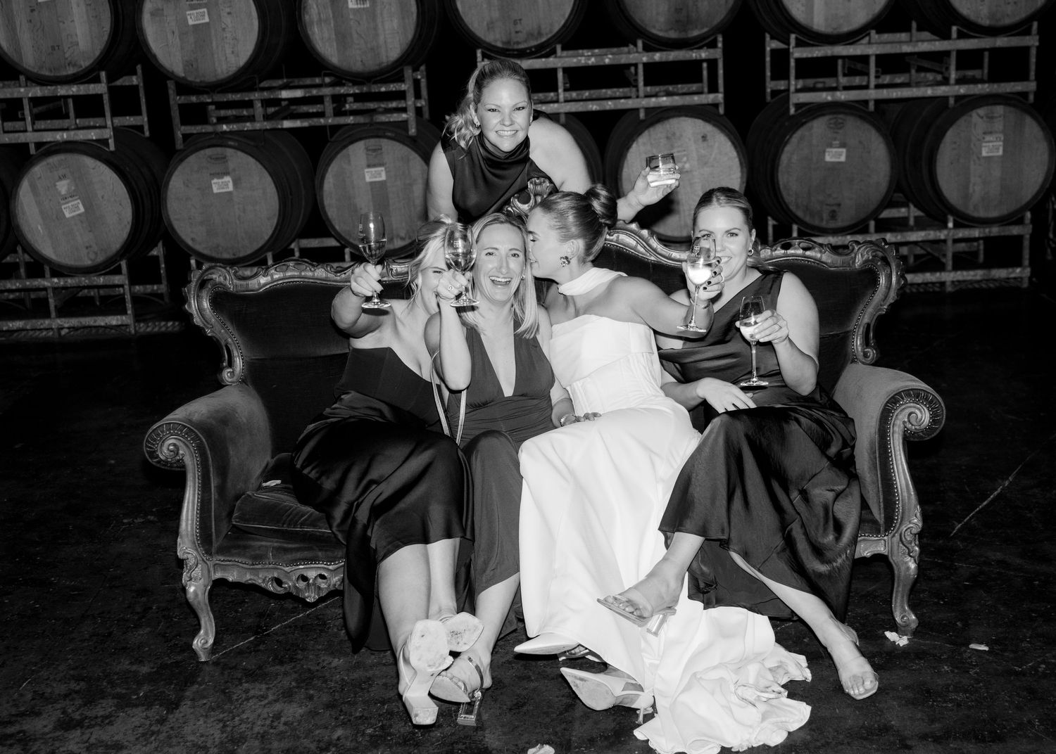 Bride and bridesmaids in black dresses laughing on a sofa in front of wine barrels at a winery wedding.