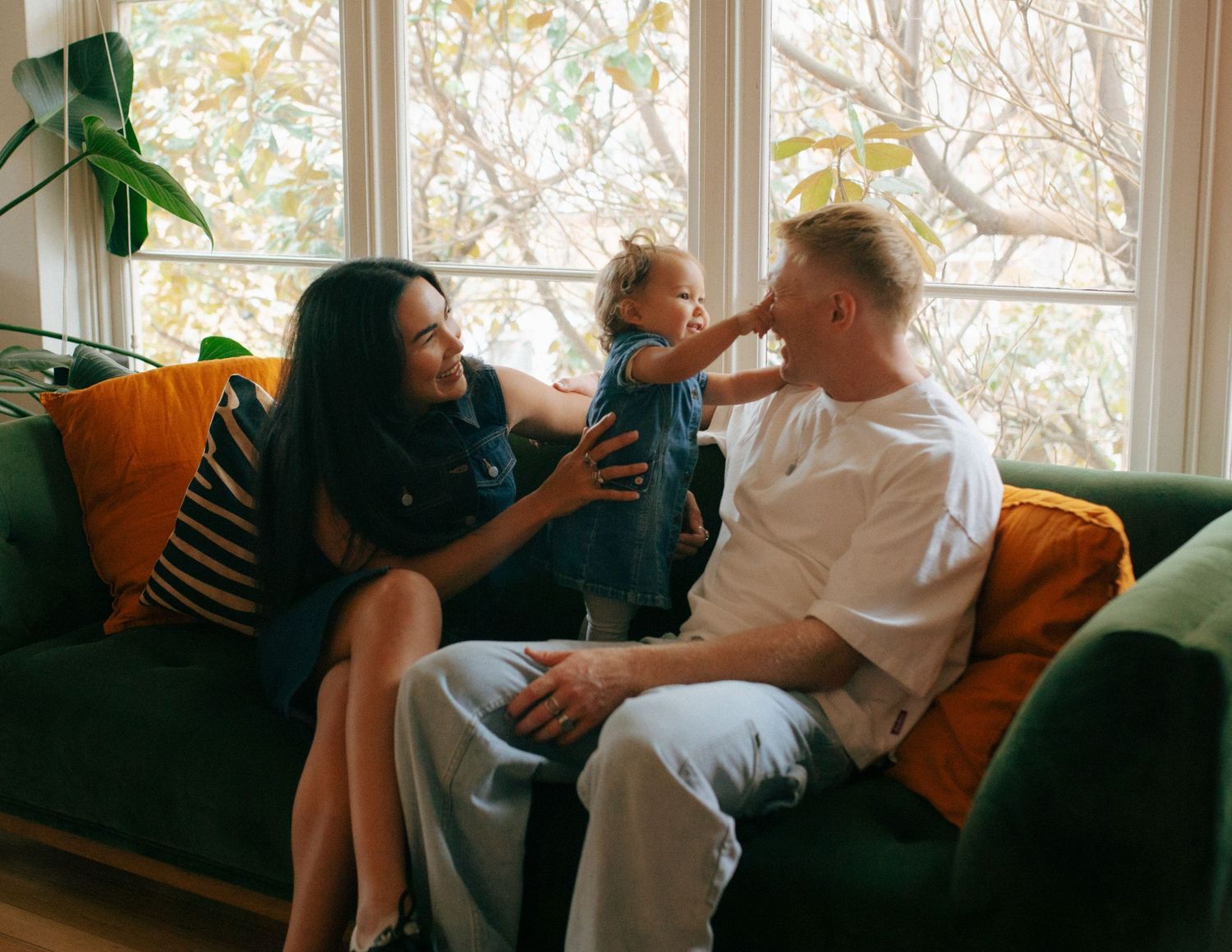 Happy family with toddler playing on green couch in sunlit living room with orange cushions.
