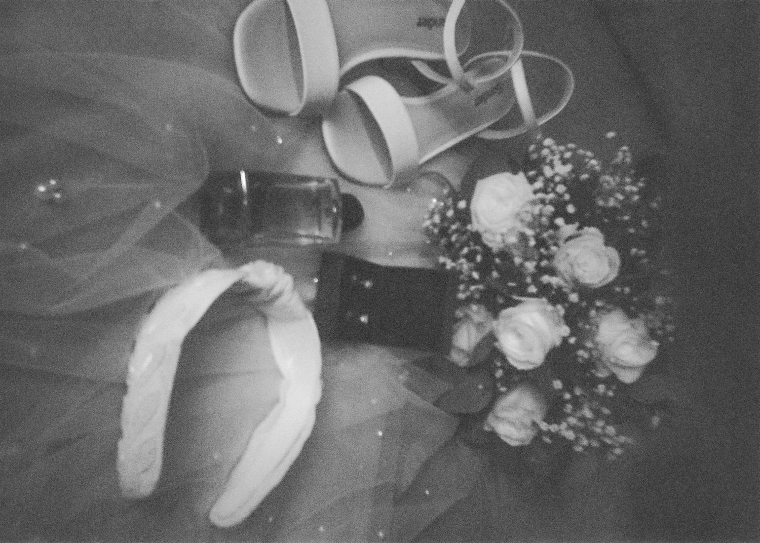 Black and white flat lay of bridal accessories including white roses bouquet, heels, perfume, and a hair bow on tulle.