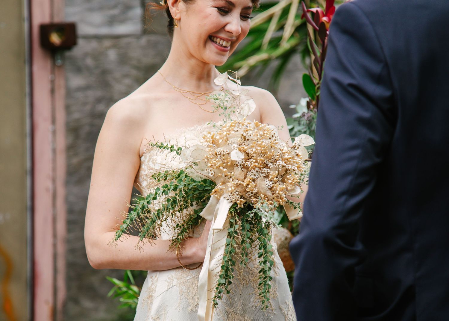 Smiling bride in strapless lace gown holding a dried floral bouquet at ceremony.