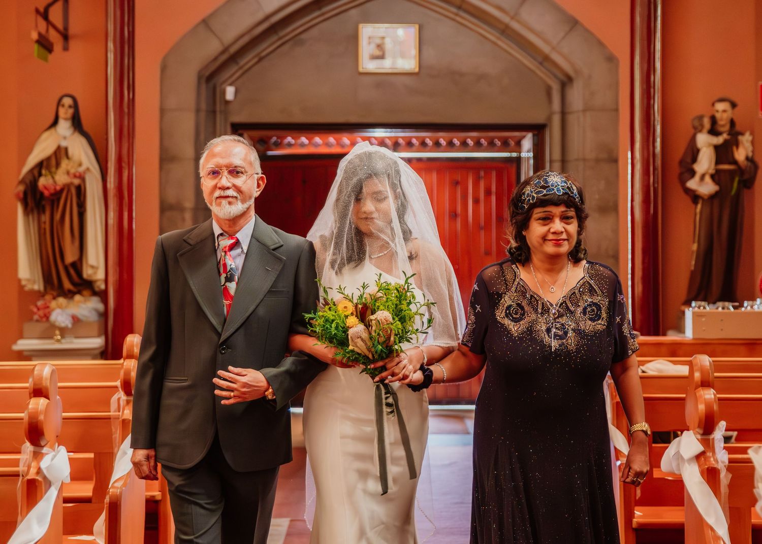 Bride walks down church aisle with parents during traditional wedding ceremony procession.