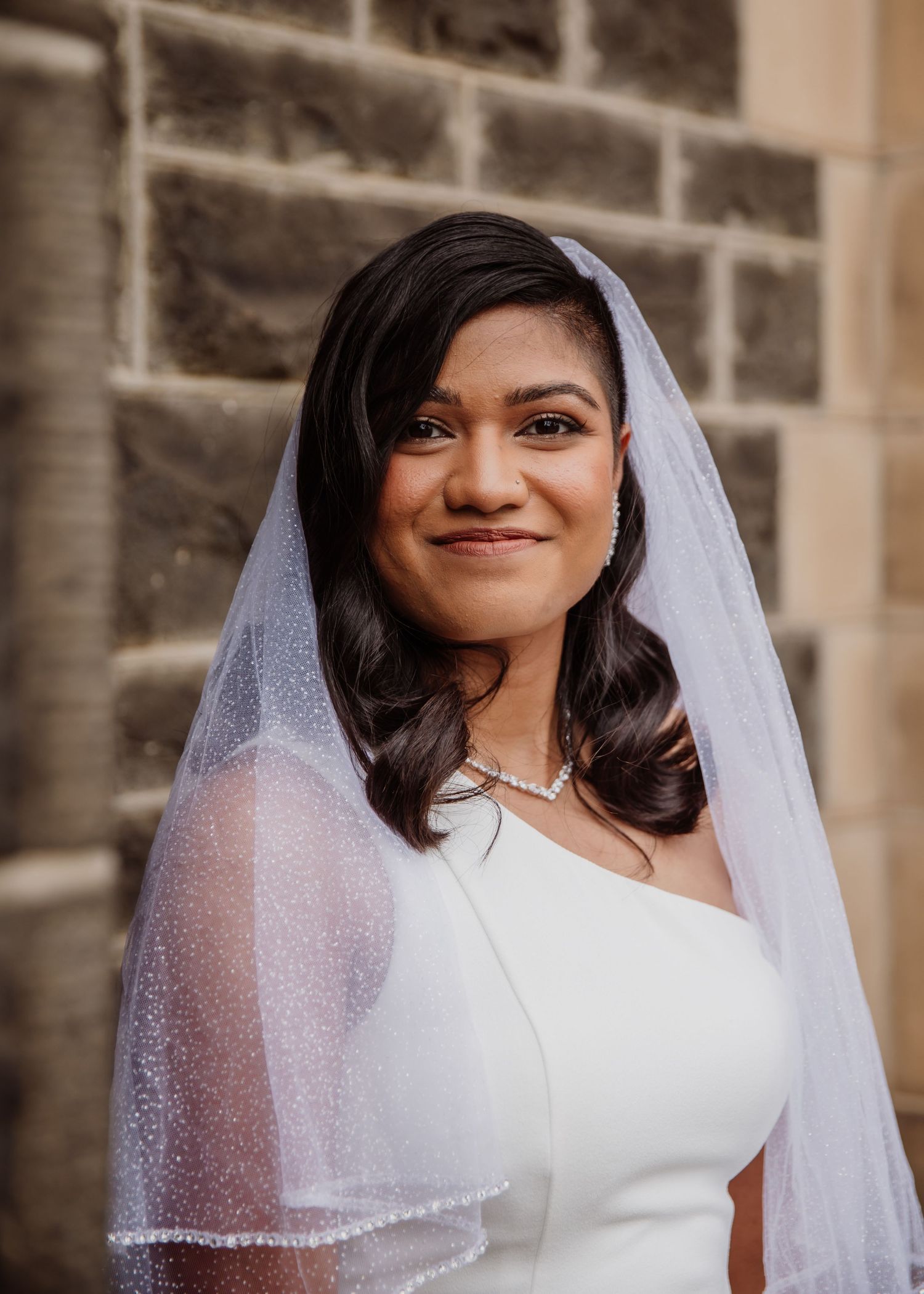 Beautiful bride wearing a white gown, veil, and diamond necklace poses in front of a stone church wall.