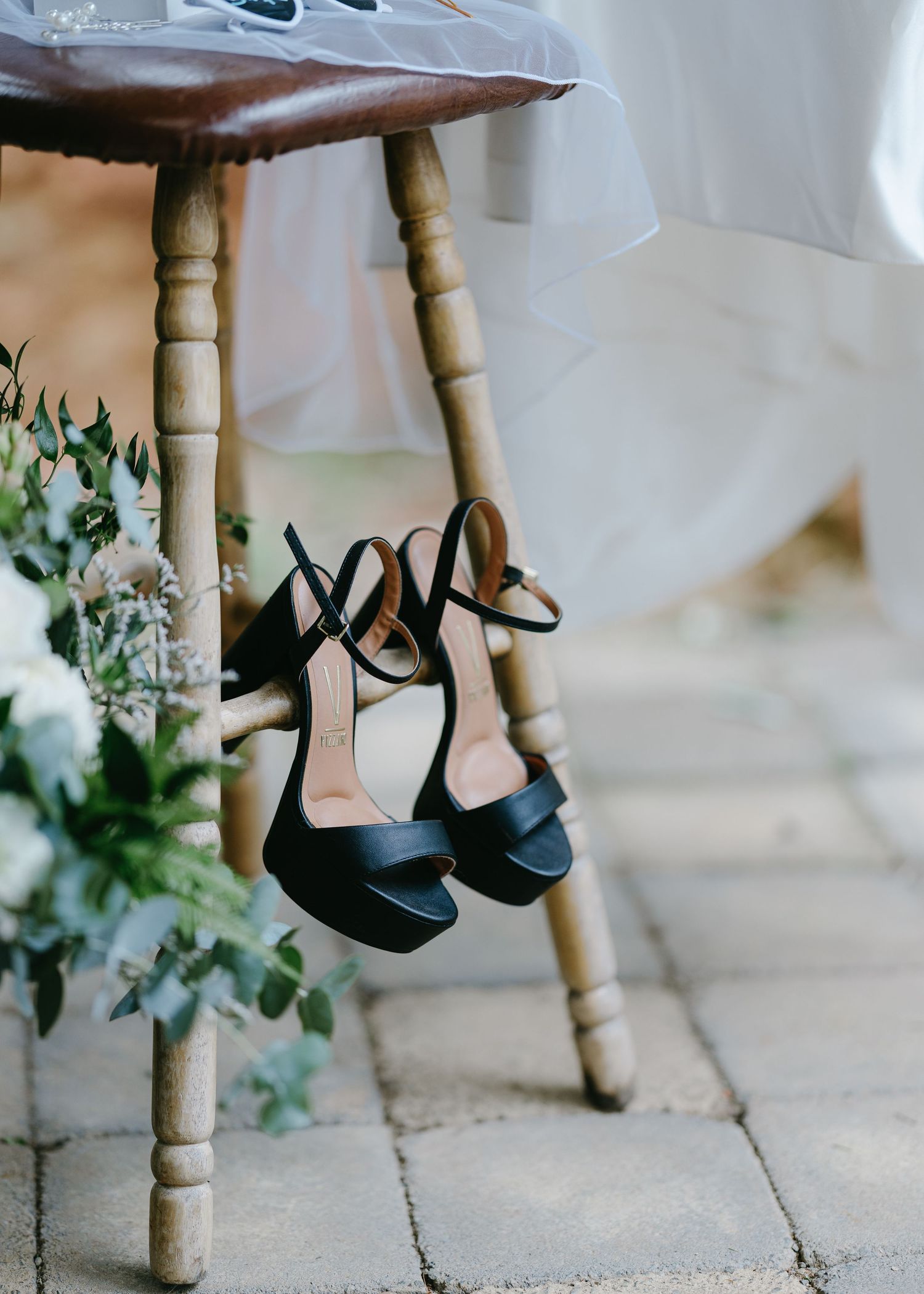 Black strappy high heel wedding shoes hanging from a wooden stool on a stone patio with white flowers.