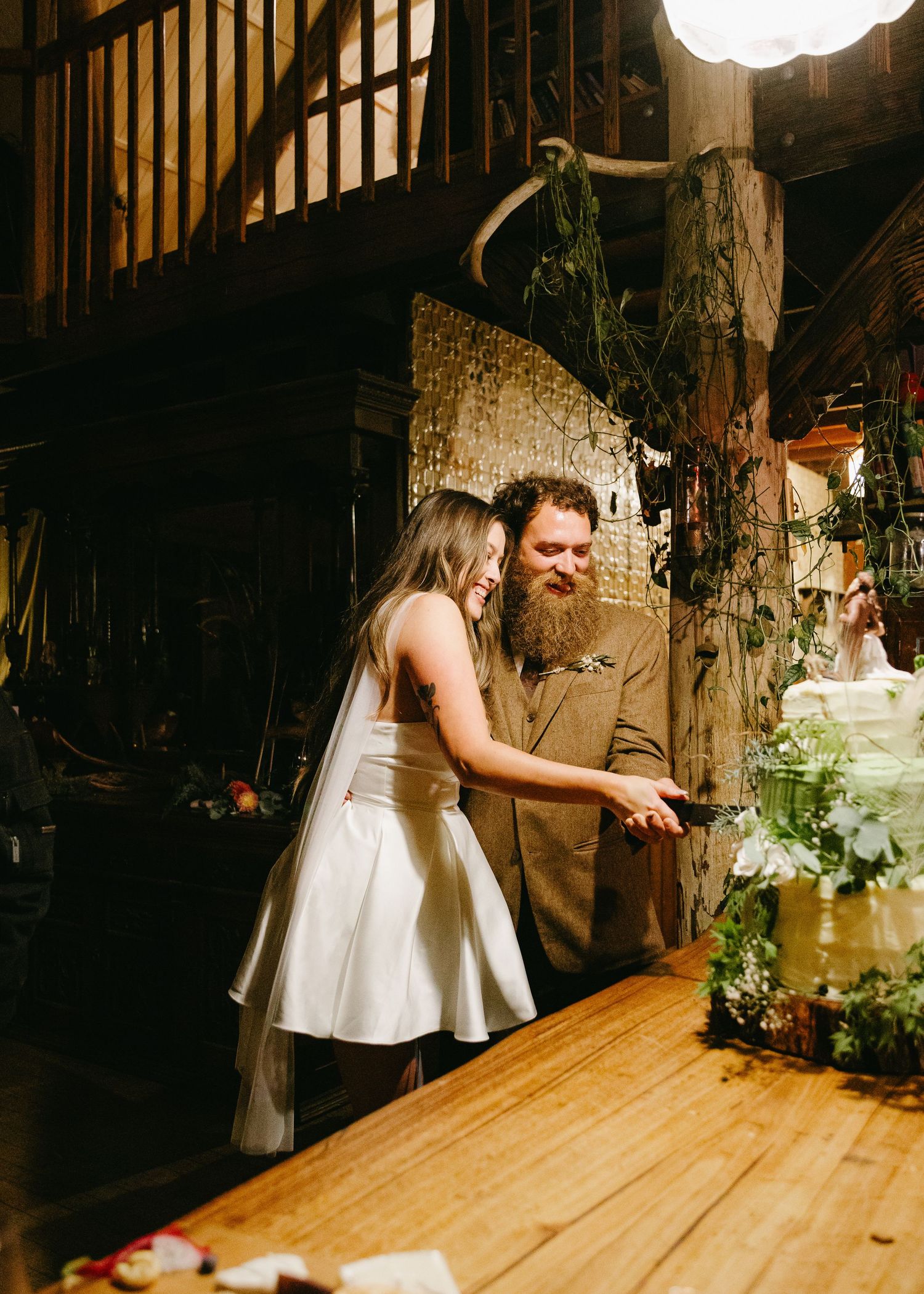 Bride and groom cutting wedding cake together in a rustic barn venue with warm lighting.