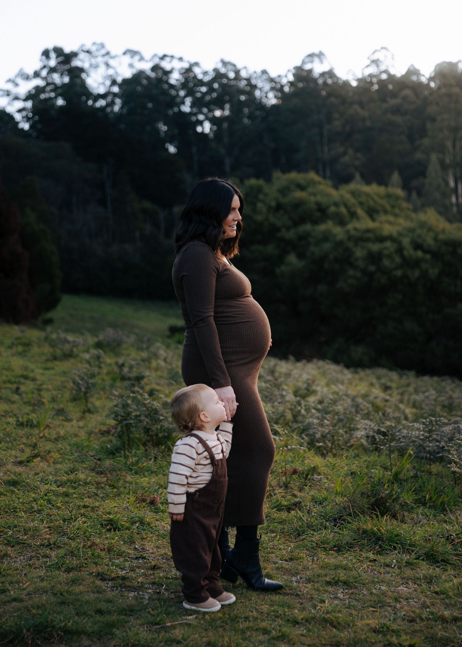 Pregnant woman in brown dress standing outdoors with toddler clinging to her leg at golden hour.