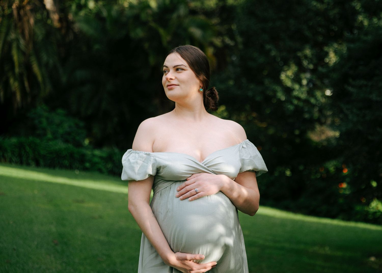 Pregnant woman in sage green off-shoulder maternity gown standing in lush garden, cradling baby bump.