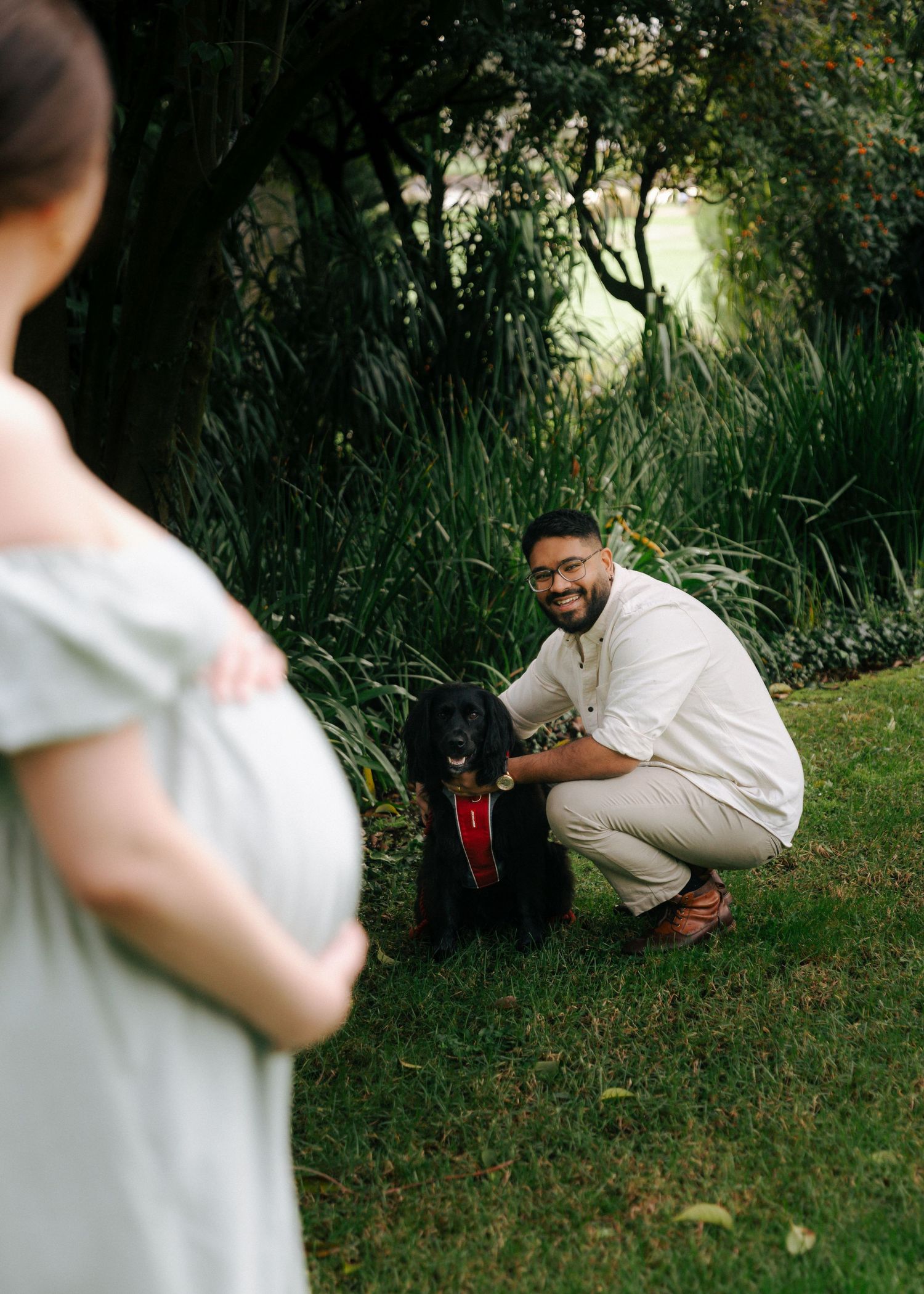 Smiling bearded man in white shirt posing with black dog in lush garden during maternity shoot.