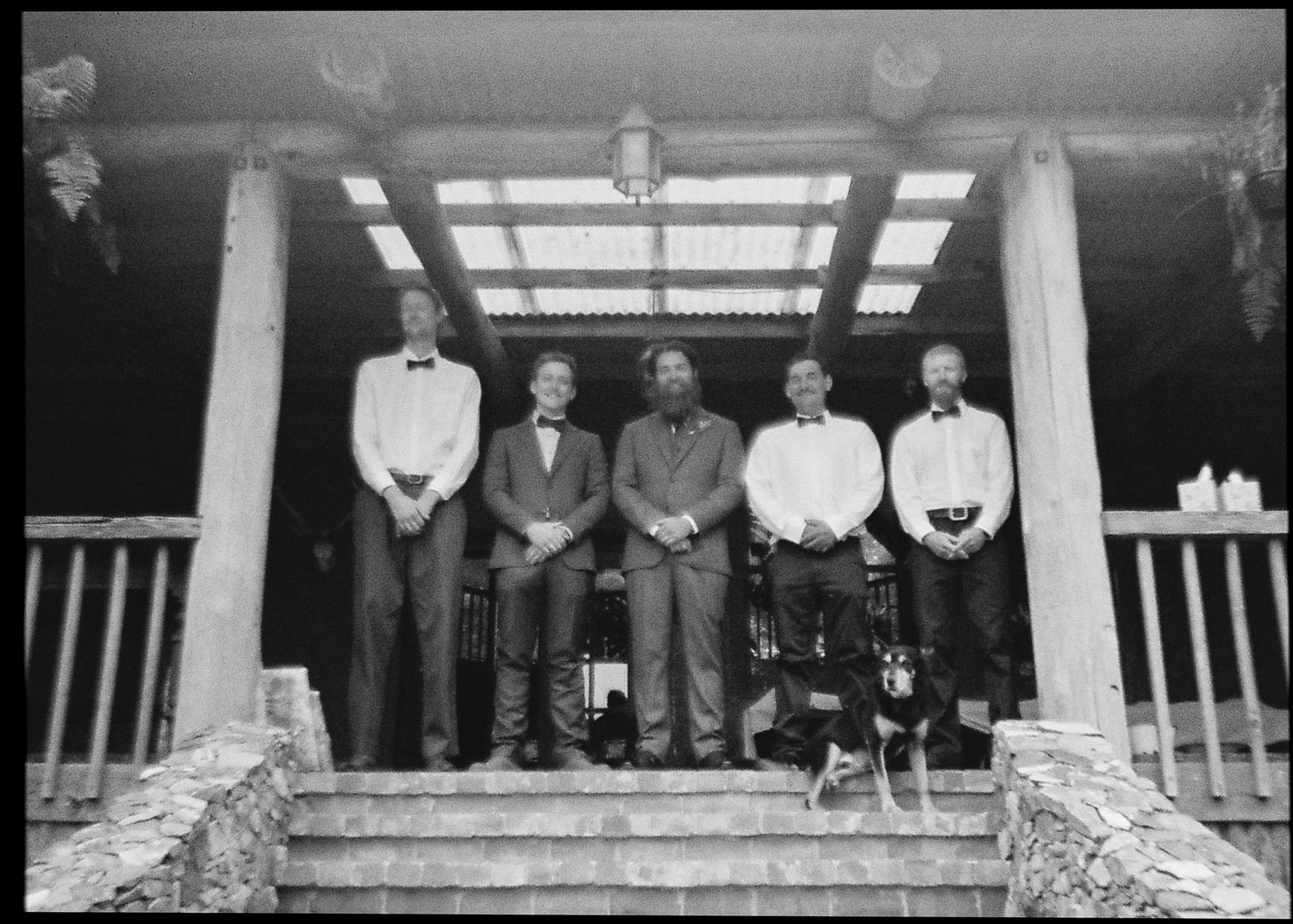 Black and white photo of five formally dressed men posing on steps of a wooden porch at a wedding.