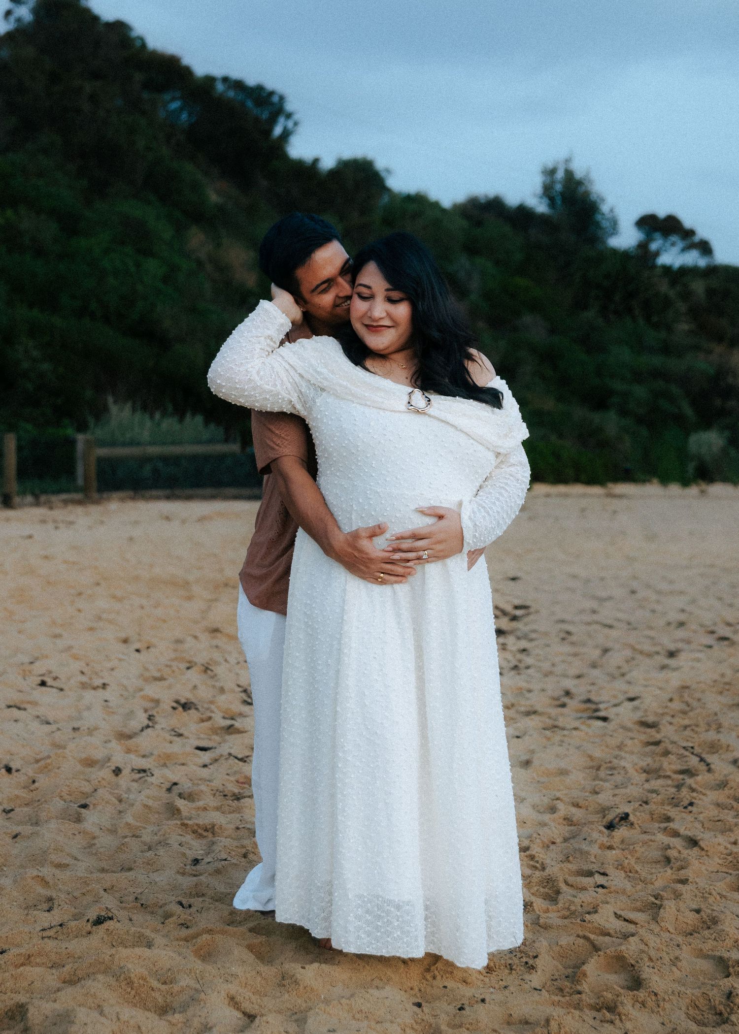 Couple maternity portrait on sandy beach, man kissing pregnant woman in white lace gown.