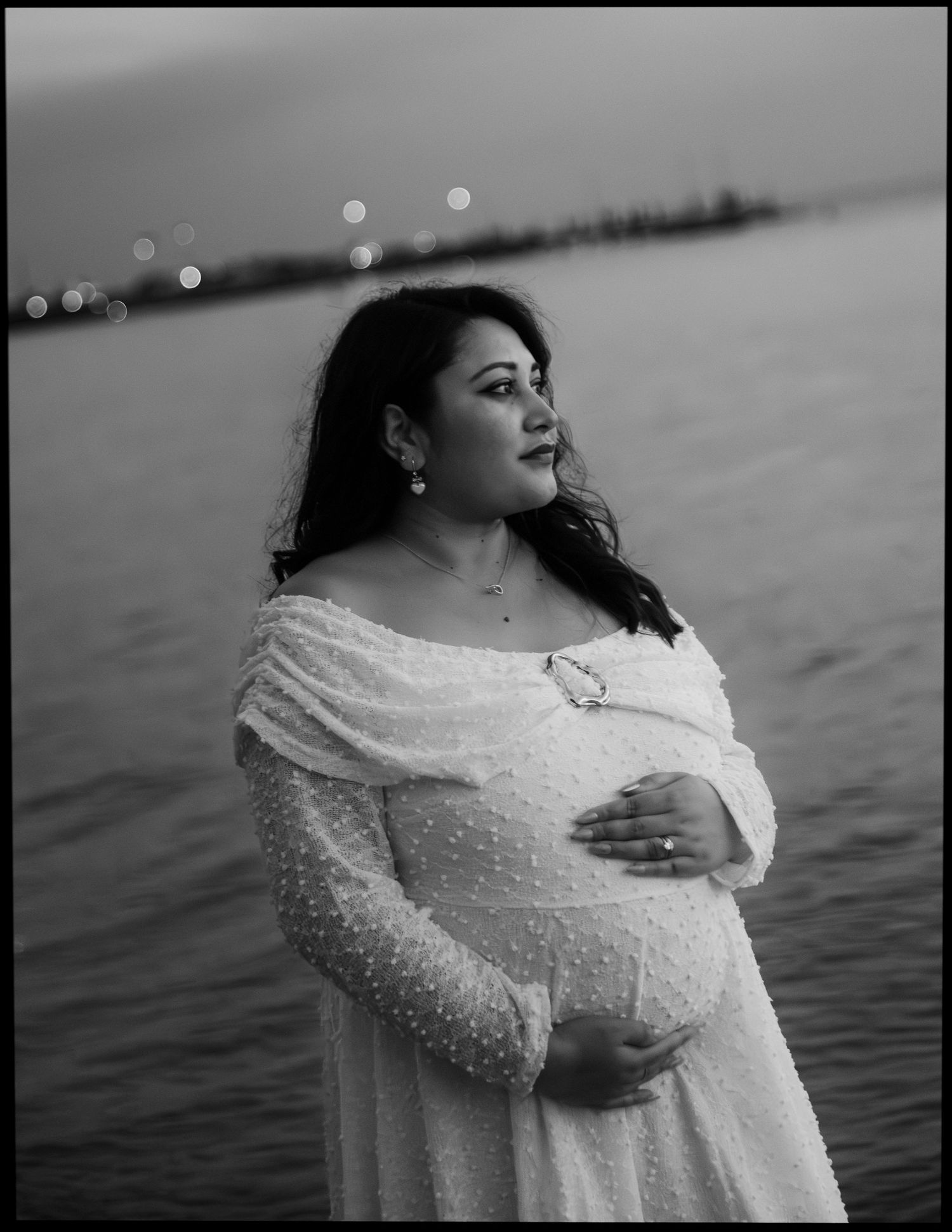 Black and white maternity portrait of pregnant woman in white lace dress near ocean at dusk.