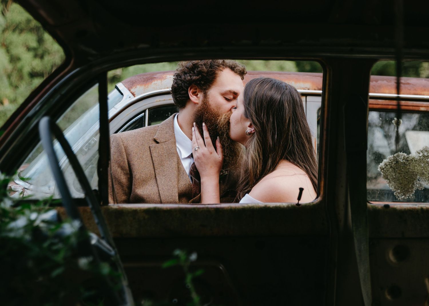 Bride and groom share a romantic kiss inside a vintage rusty car.