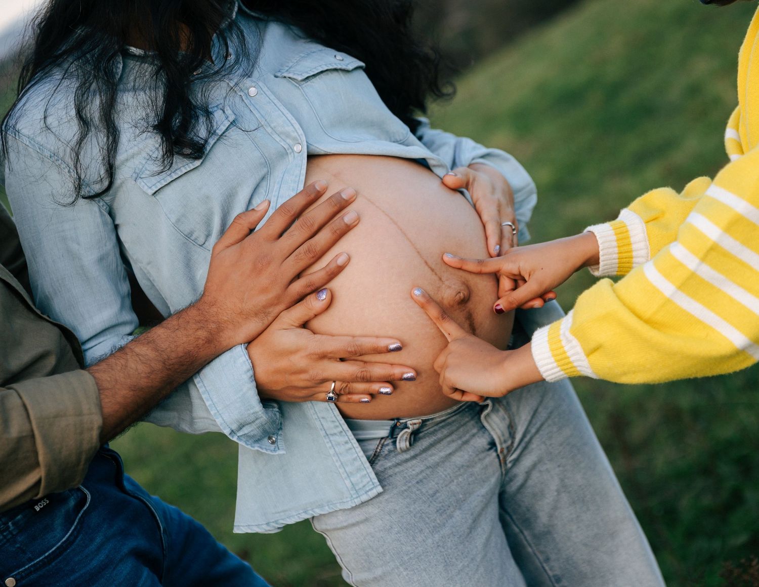Close-up of hands tenderly touching a pregnant belly outdoors, with a child in yellow sweater joining in.