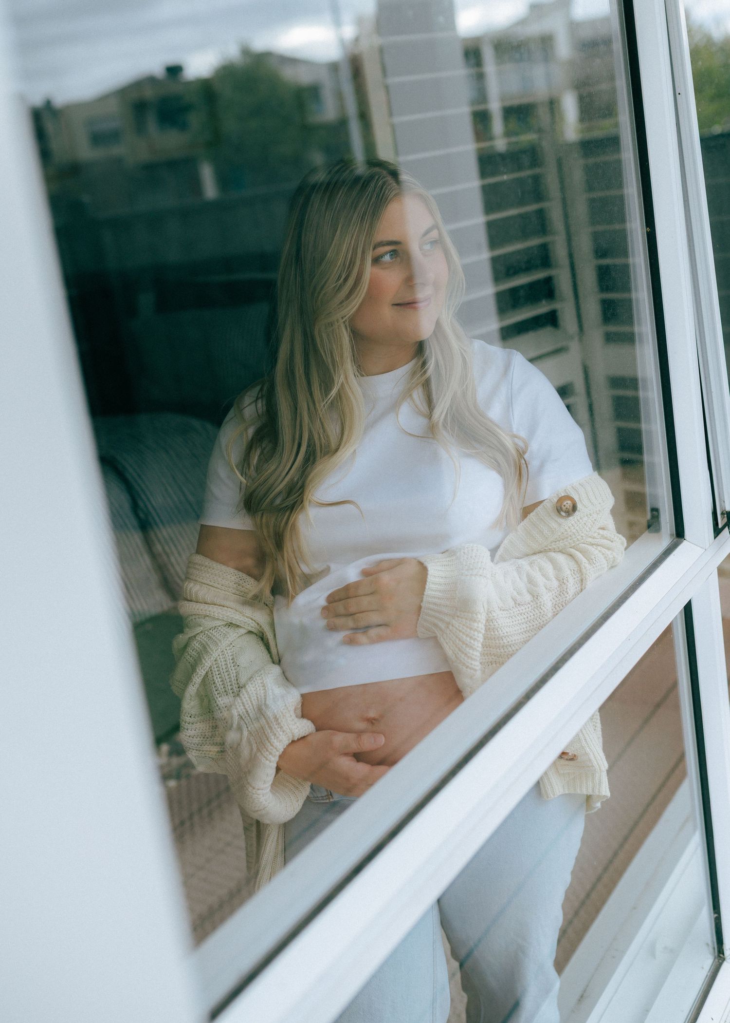 Pregnant woman in white top and cream cardigan cradling baby bump while gazing through window.