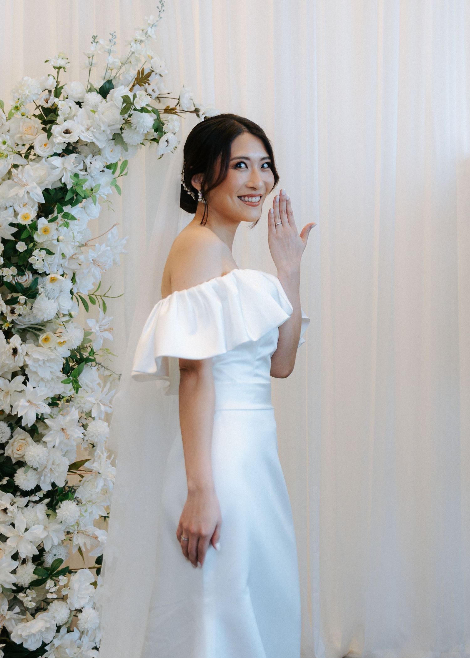 Radiant bride in ruffled off-shoulder white gown posing beside white floral arch at ceremony.