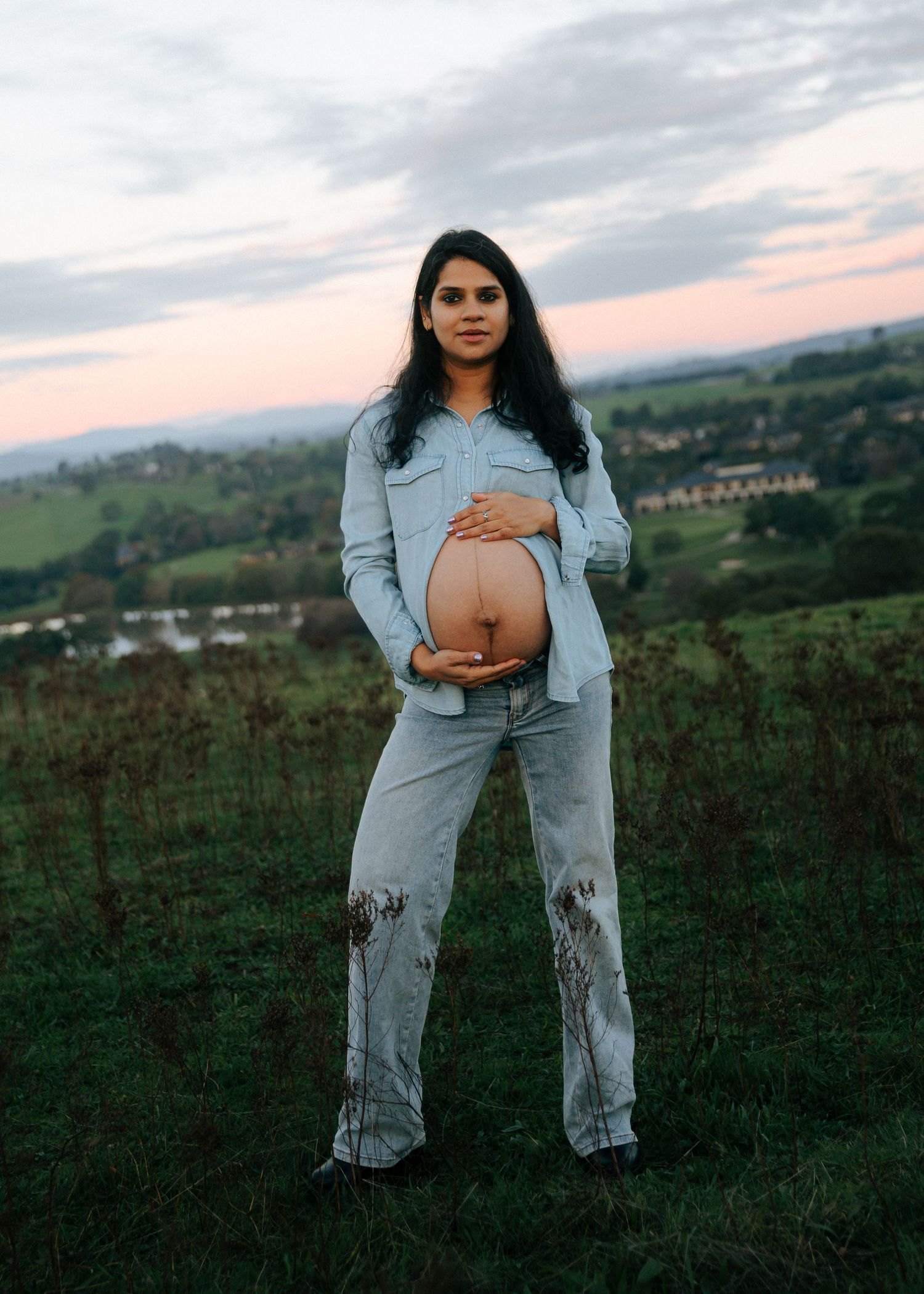 Confident pregnant woman in denim outfit cradling her bare belly on a green hill at sunset.