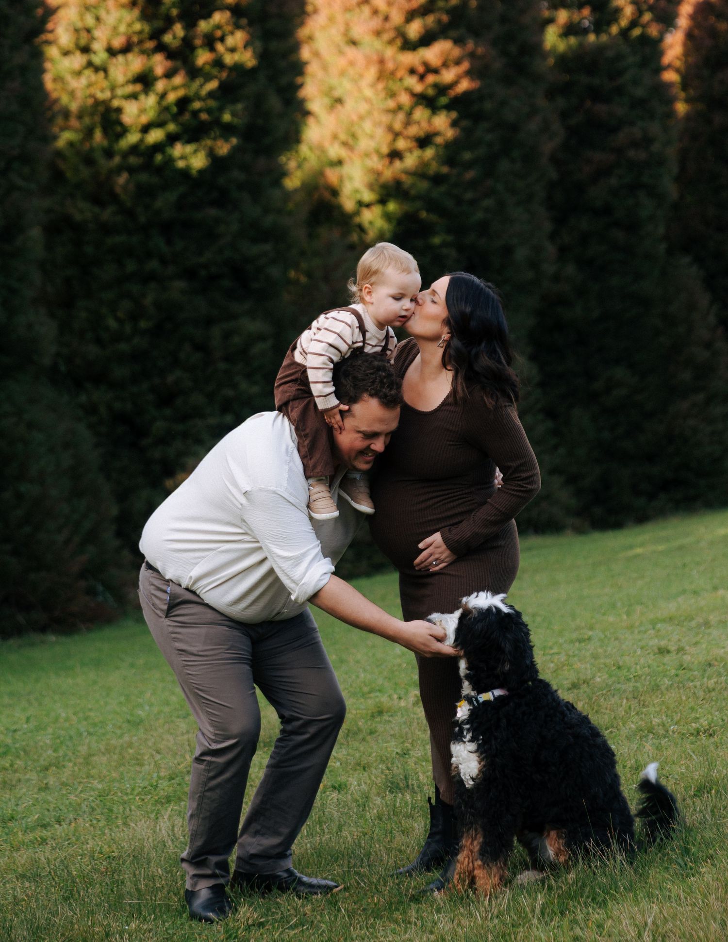 Family moment with dad lifting baby toward pregnant mom while dog sits nearby.